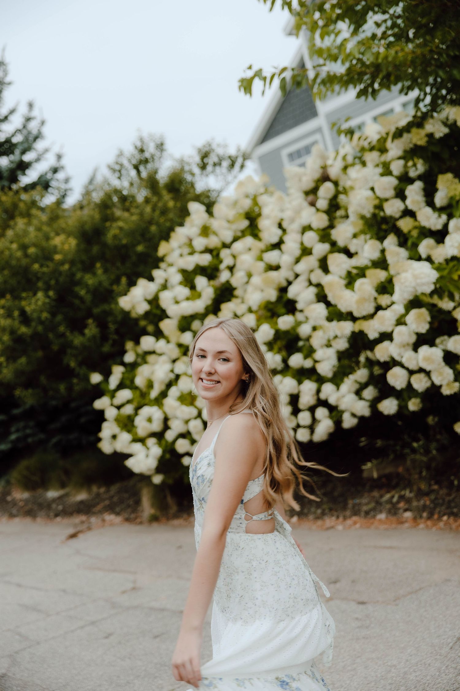 Young blonde woman in elegant white embroidered dress smiling outdoors with blooming white hydrangeas in background.