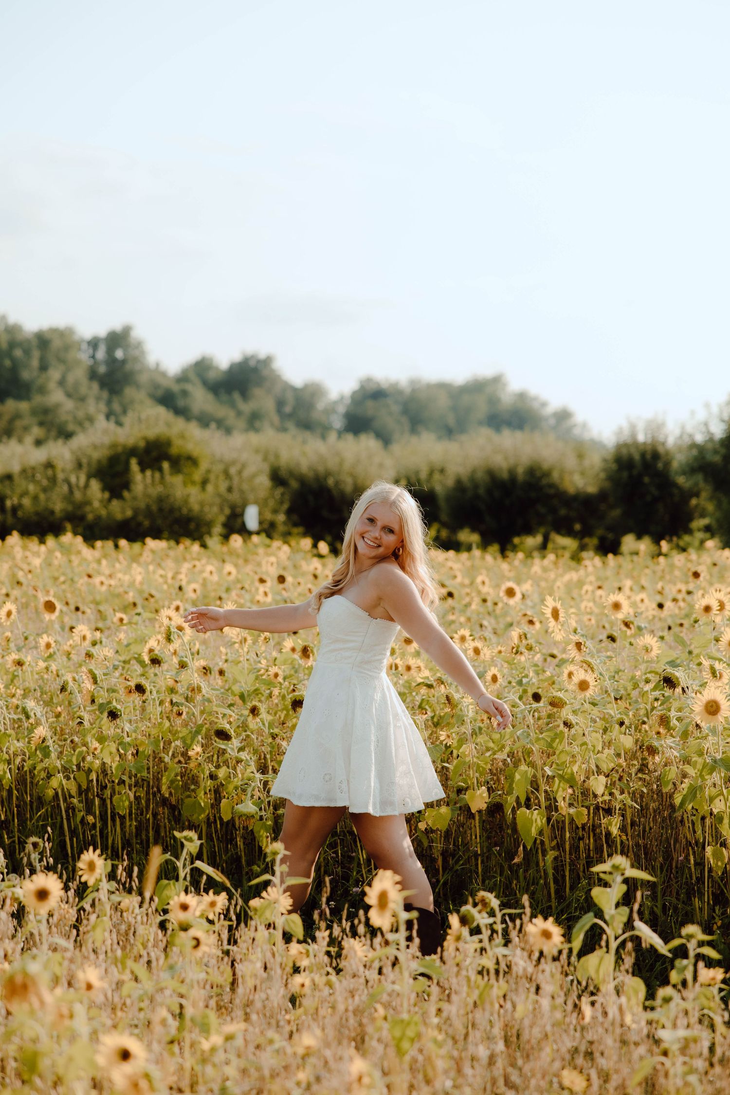Young blonde woman in white sundress twirling joyfully in a golden sunflower field on a sunny day.