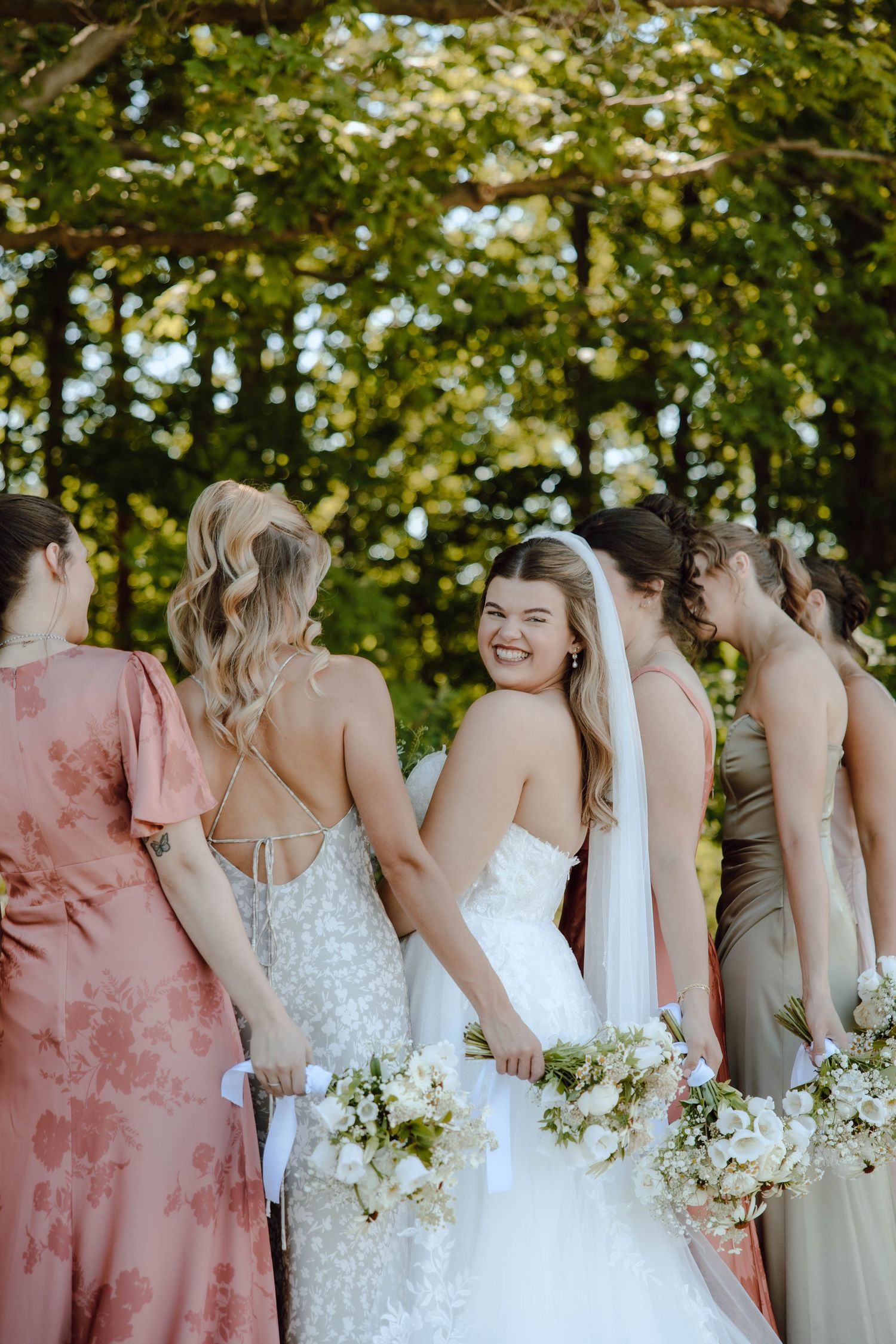 Bride in white gown laughs with bridesmaids in mixed floral dresses holding white bouquets outdoors.
