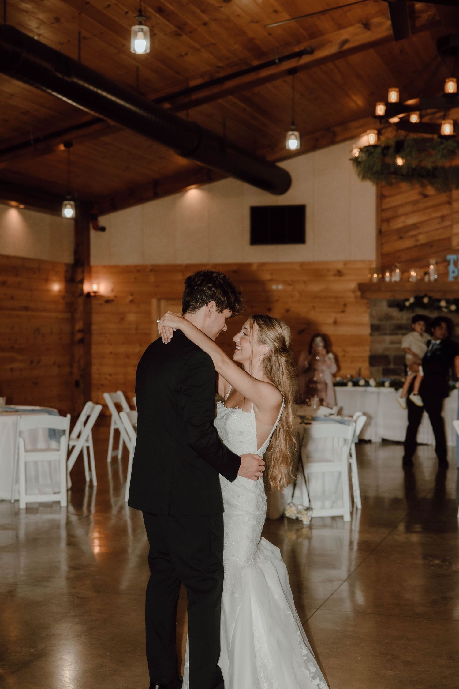 Bride and groom share their first dance in a rustic wooden reception venue with warm hanging lights.