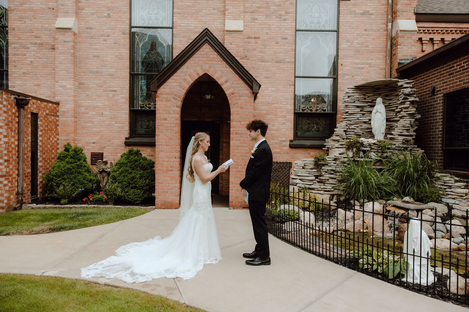 Bride and groom exchange vows outside a brick church, with her long white veil trailing on the pathway.