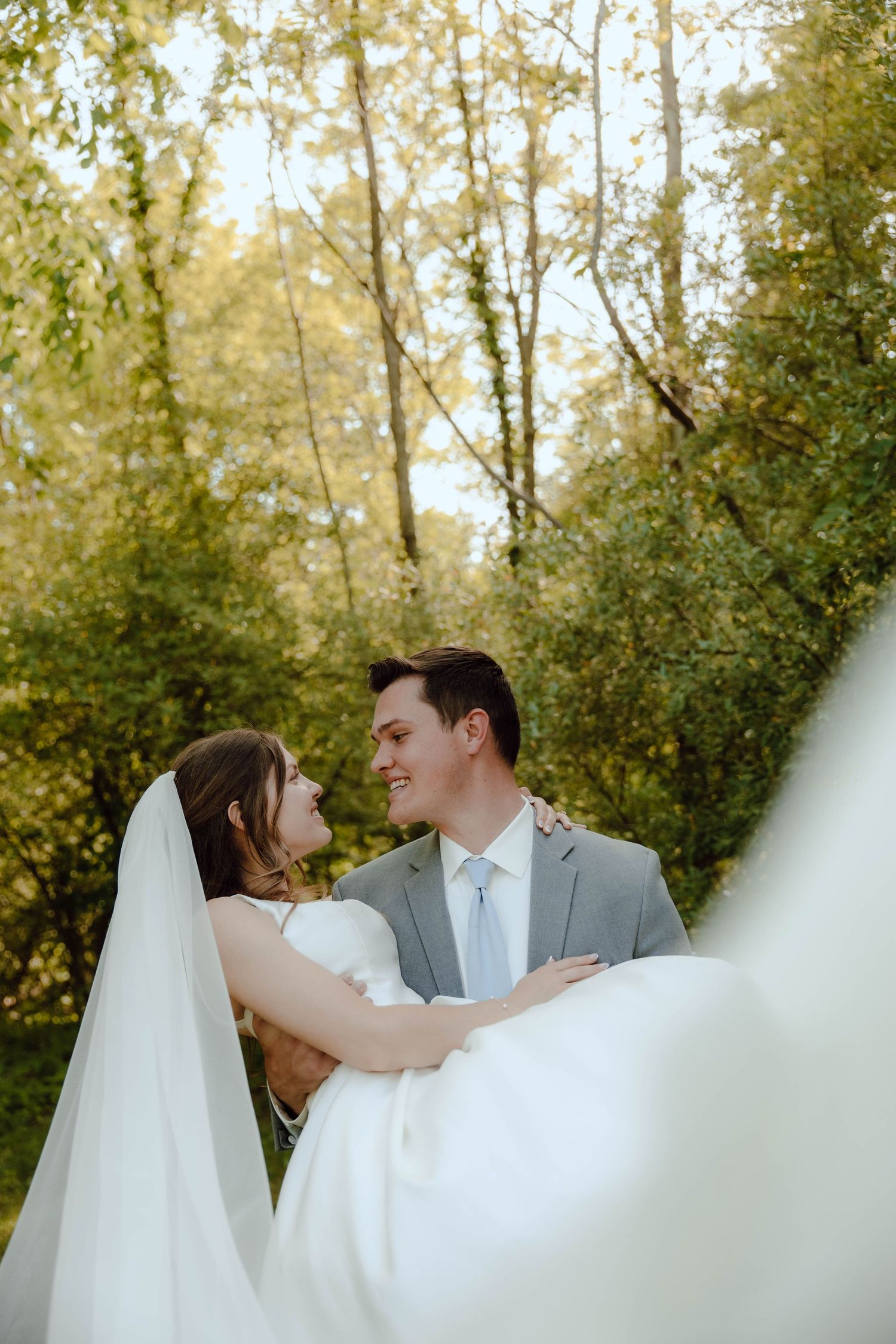 Bride and groom share a romantic moment embracing and smiling at each other outdoors among lush green trees.