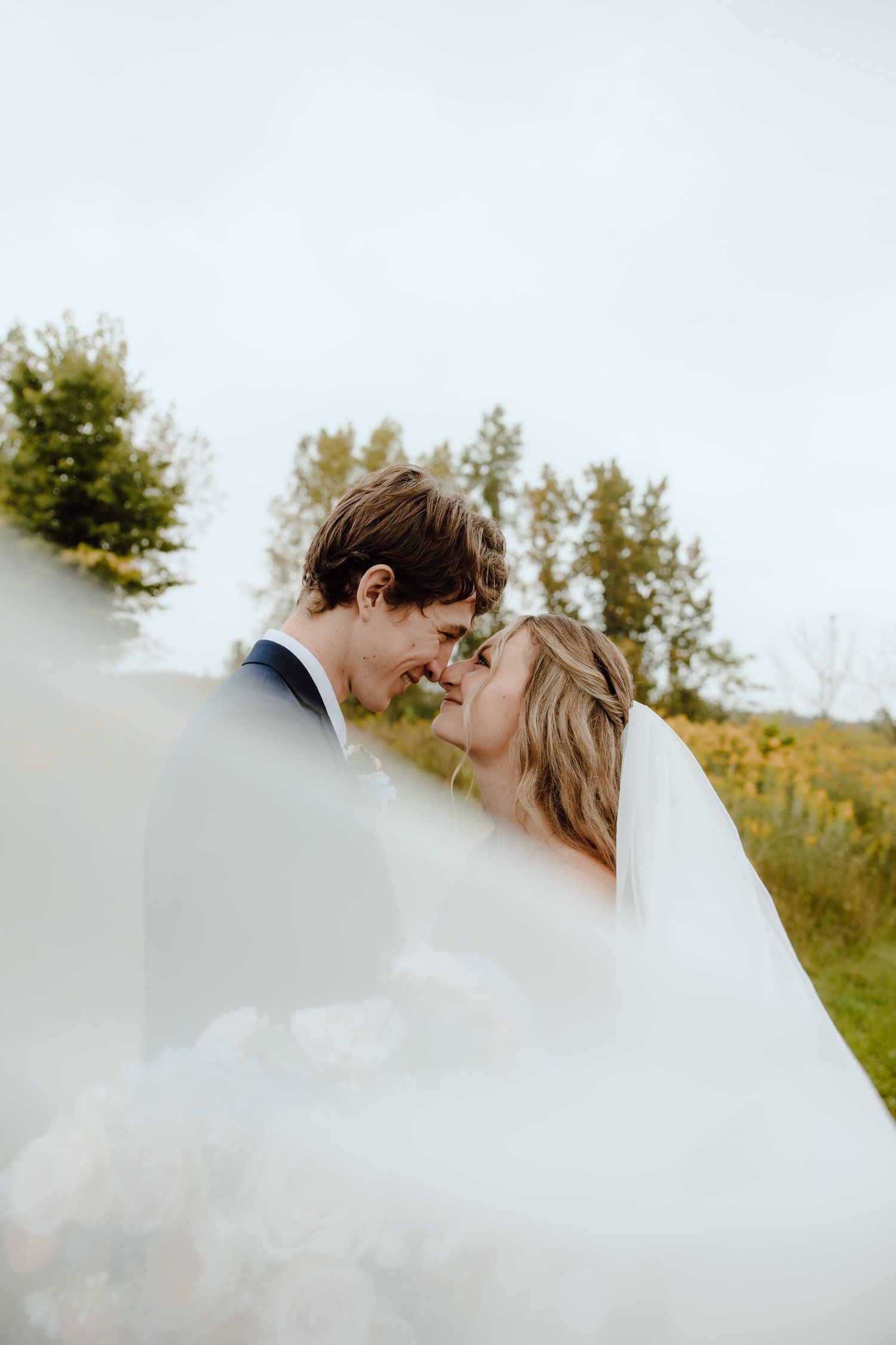 Bride and groom share a romantic almost-kiss outdoors, framed by a flowing white veil with green trees behind them.