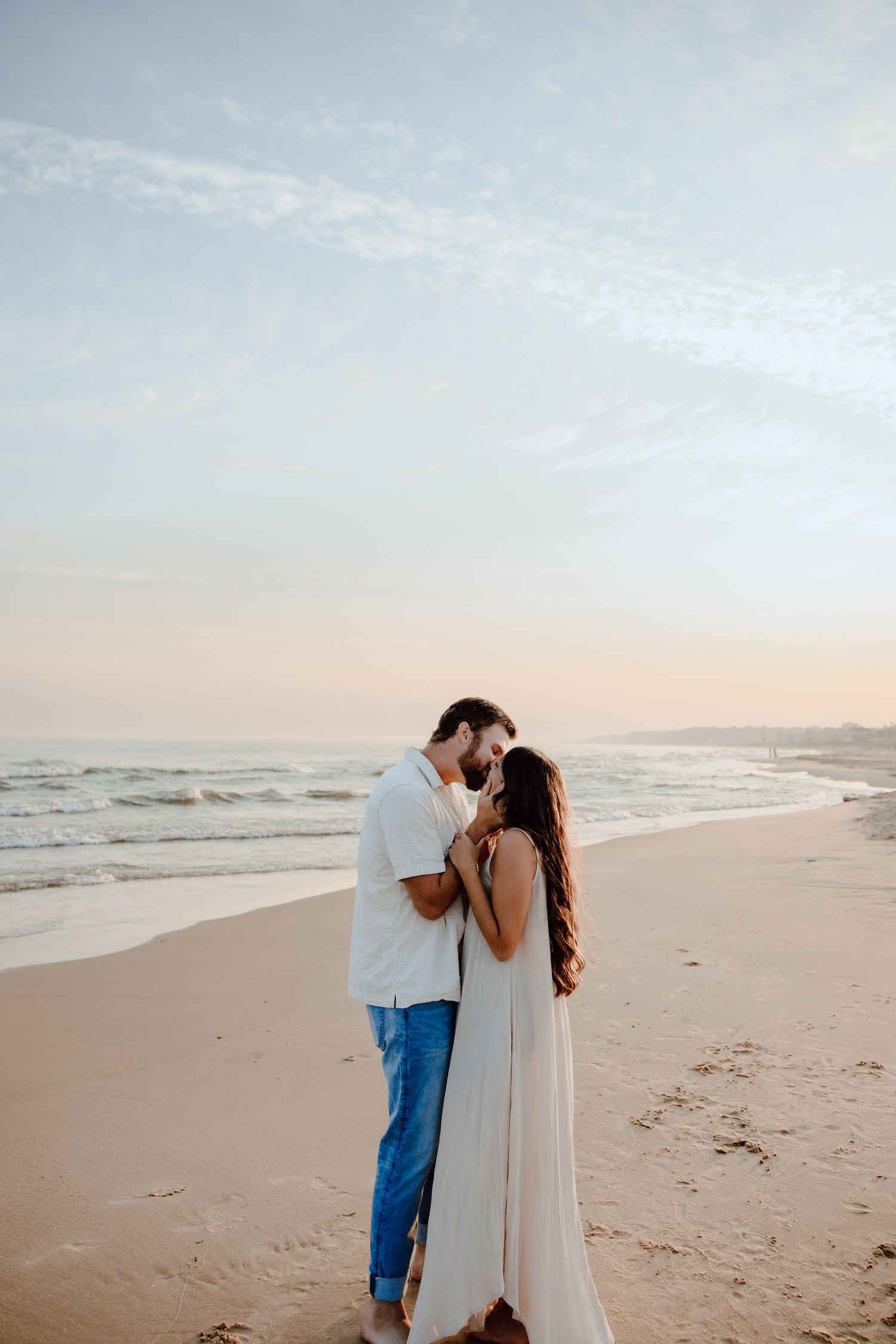 Couple in white clothing sharing tender moments while walking along an empty beach.