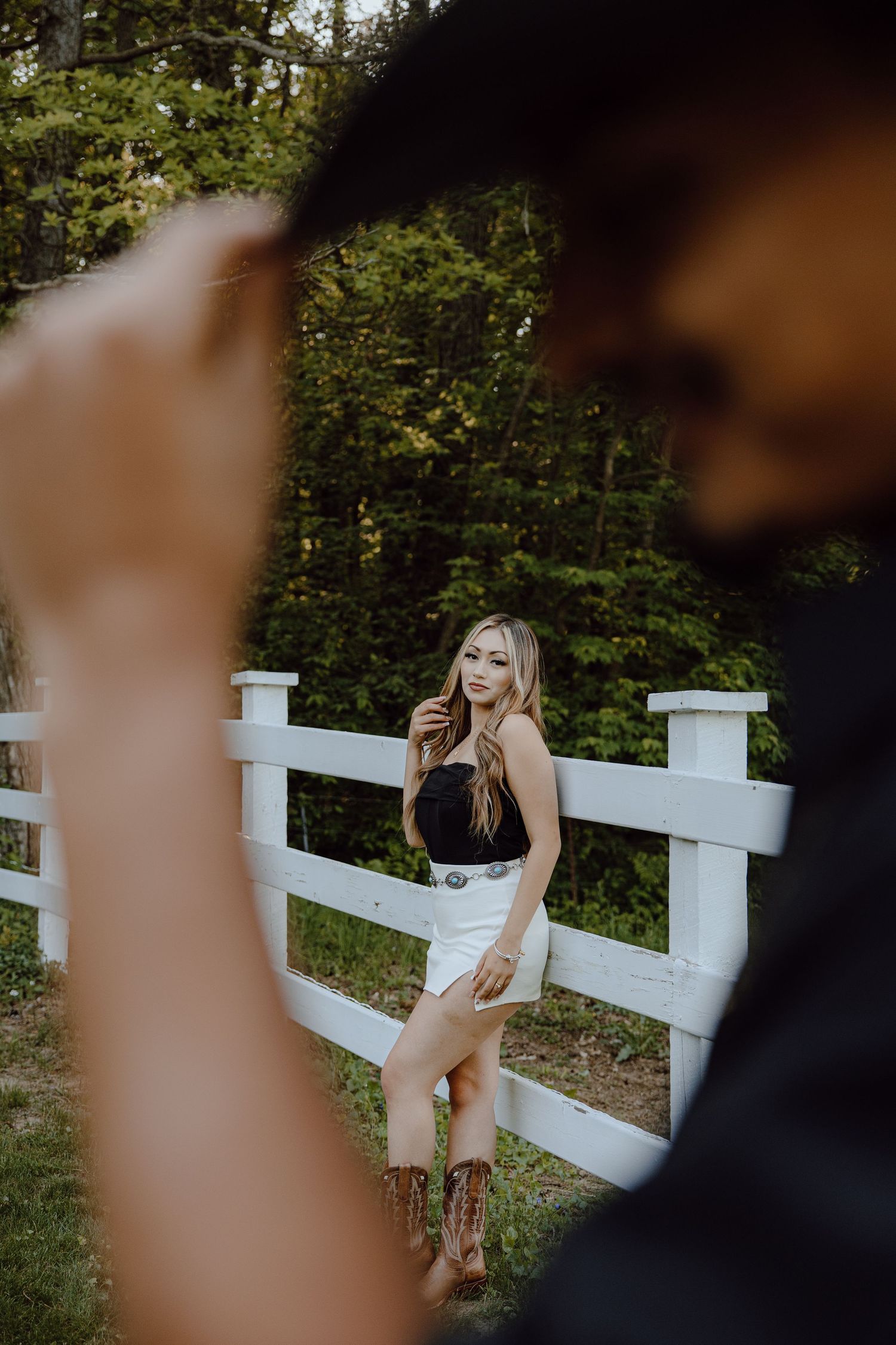 Artistic photo through foreground elements of someone posing by a white fence in nature.