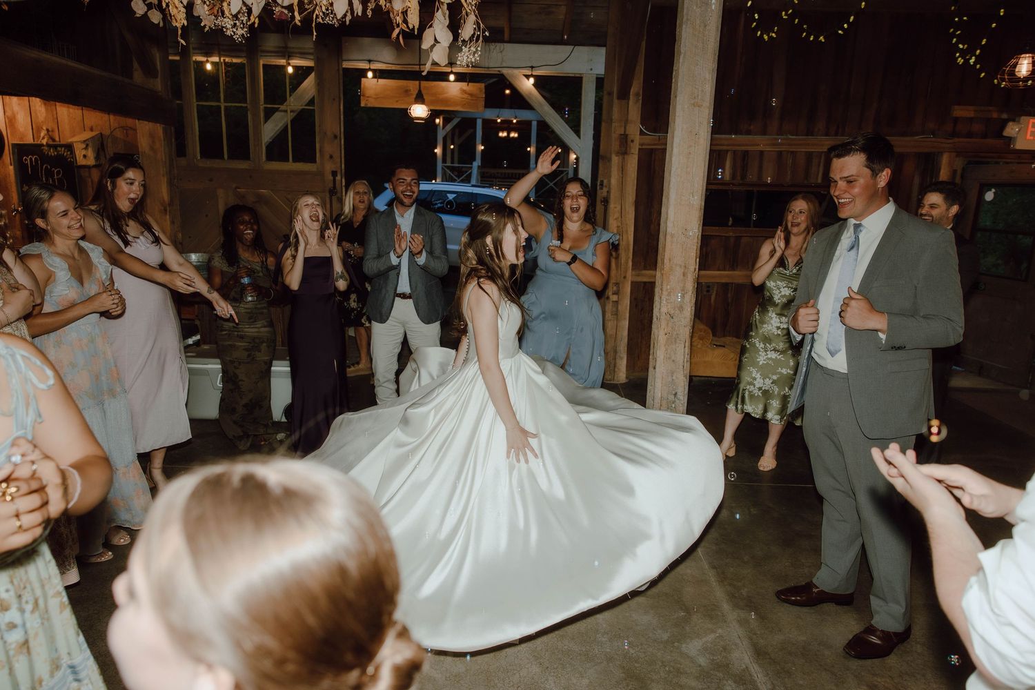 Bride twirling in white ball gown on dance floor surrounded by guests at rustic barn wedding reception.