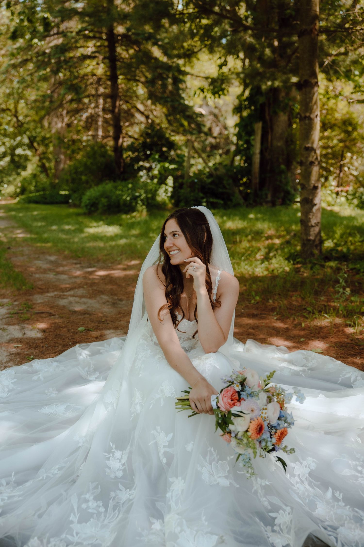 A bride in a lace gown sits outdoors holding a colorful bouquet.