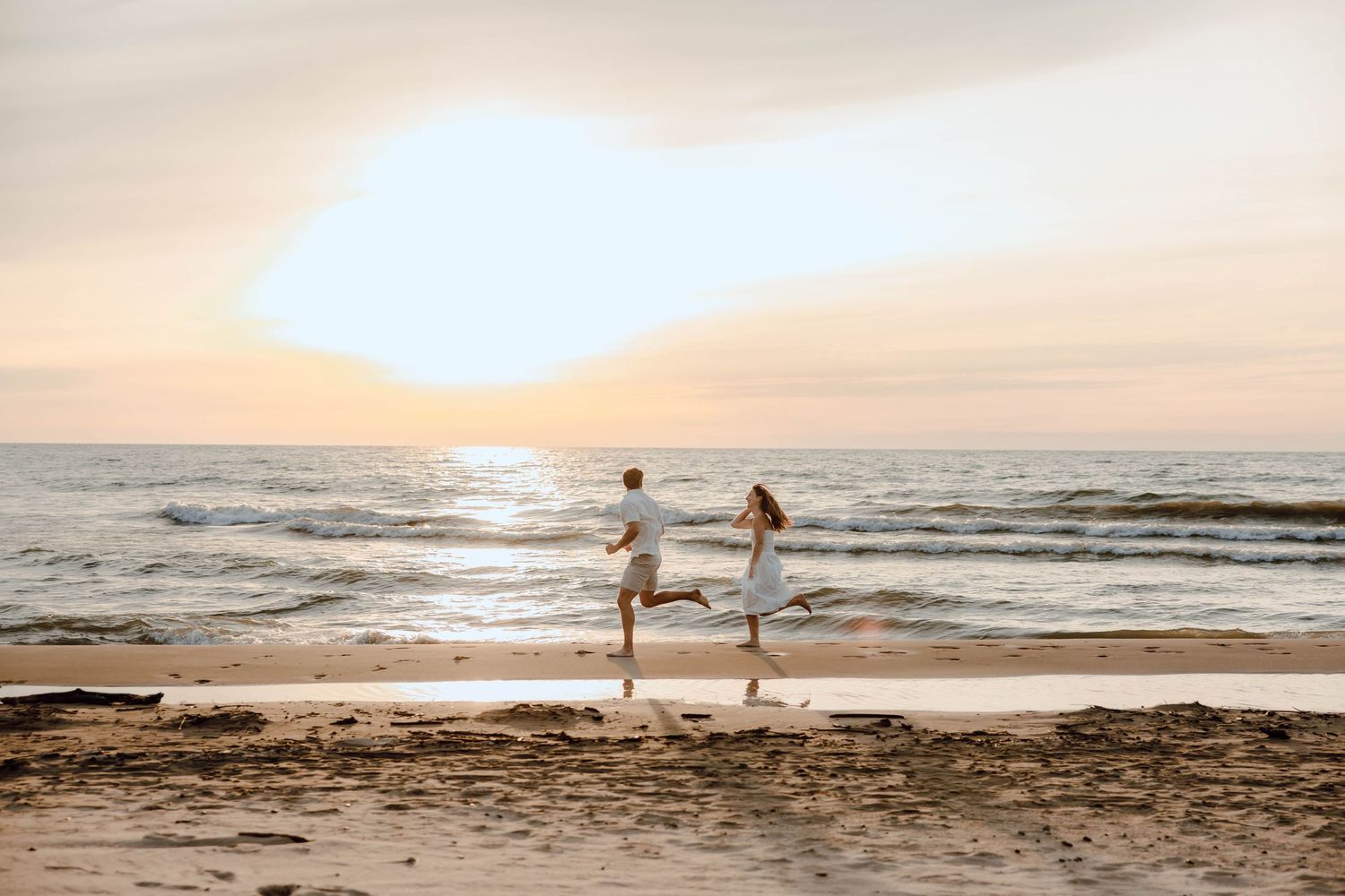 Couple holding hands and jumping joyfully on a sandy beach at sunset with ocean waves in the background.