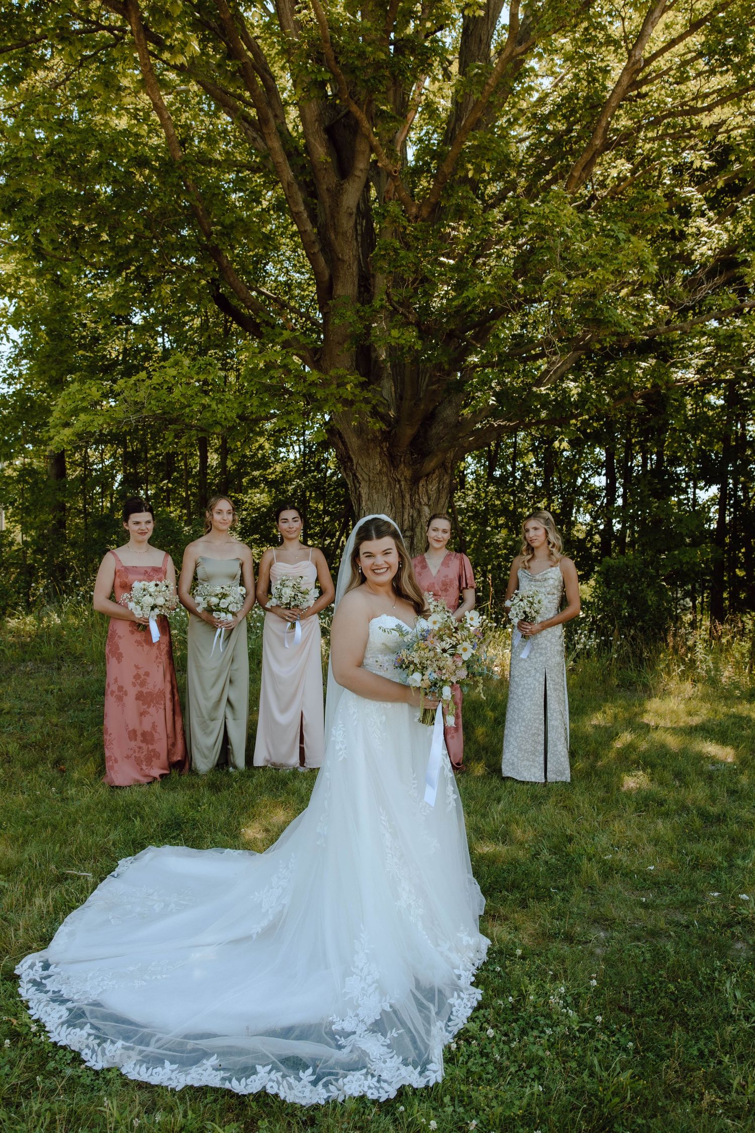 Bride in white gown with bridesmaids in pink and sage dresses holding bouquets under a large tree outdoors.