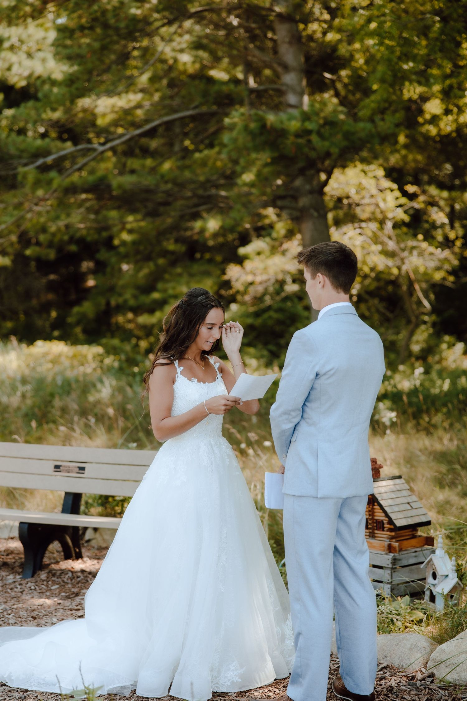 Bride in white gown wiping tears while reading vows to groom in light blue suit outdoors.