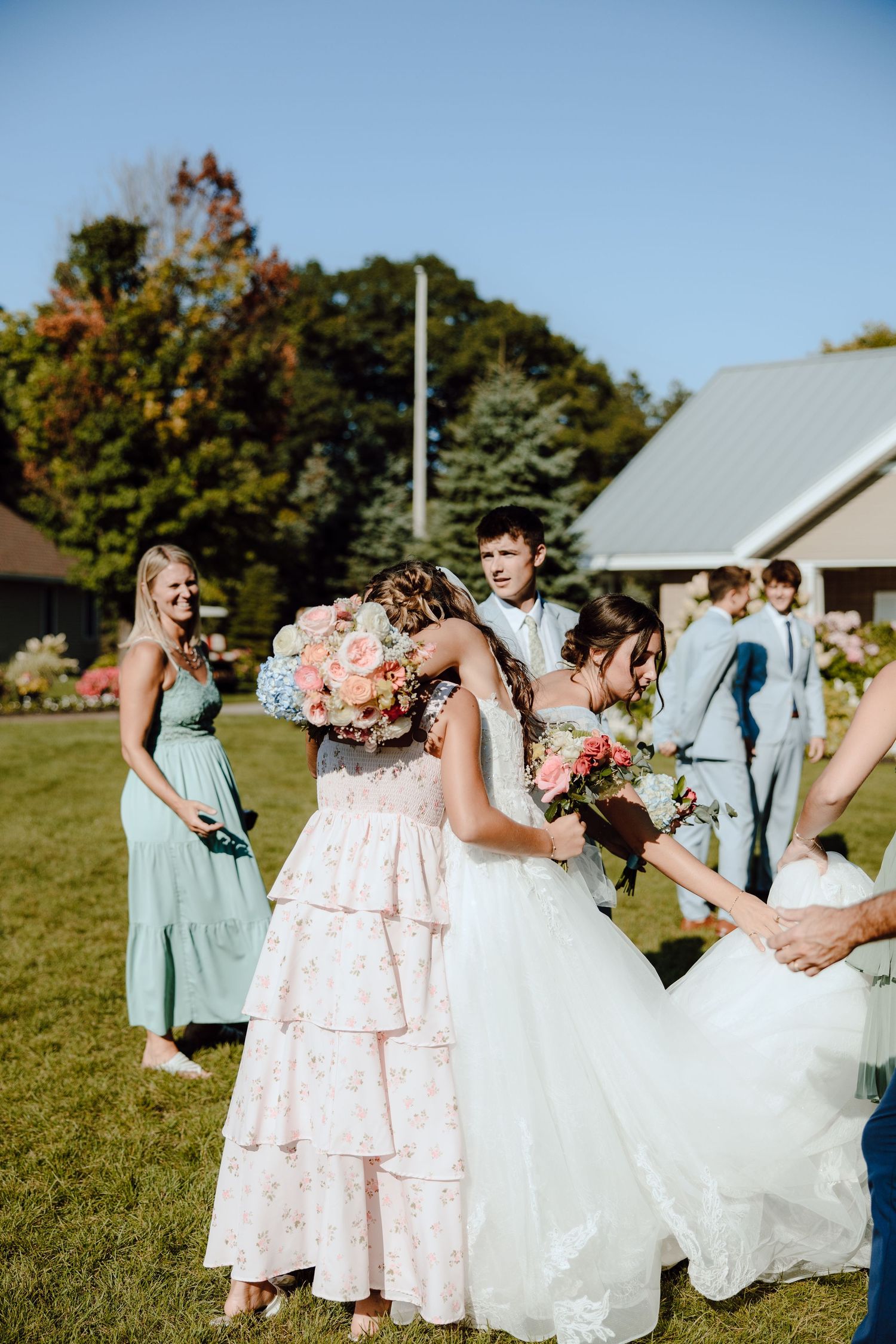 Bride in white gown embracing guest in floral dress while holding bouquet at outdoor wedding.