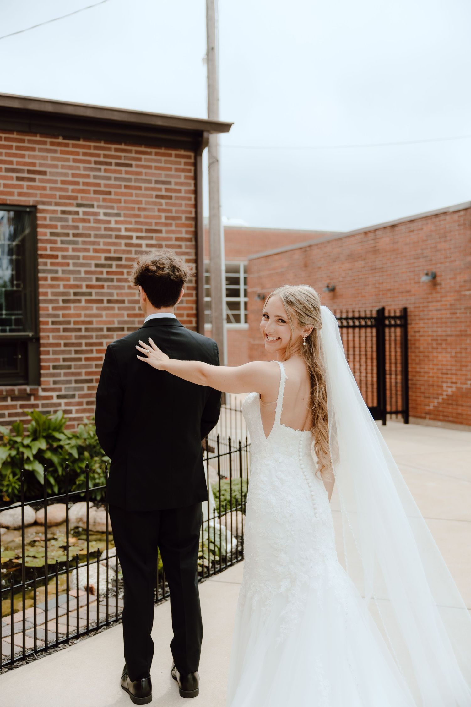 Bride in lace wedding gown taps groom's shoulder for first look outside brick church venue.