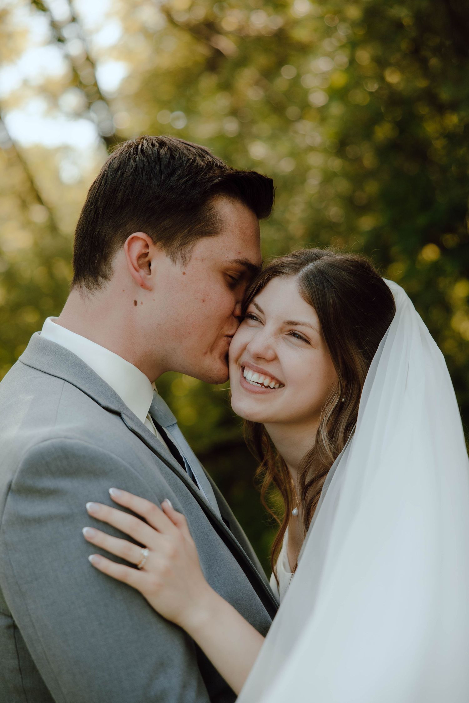 Groom tenderly kisses smiling bride on the cheek outdoors, her white veil flowing in a sunlit forest setting.