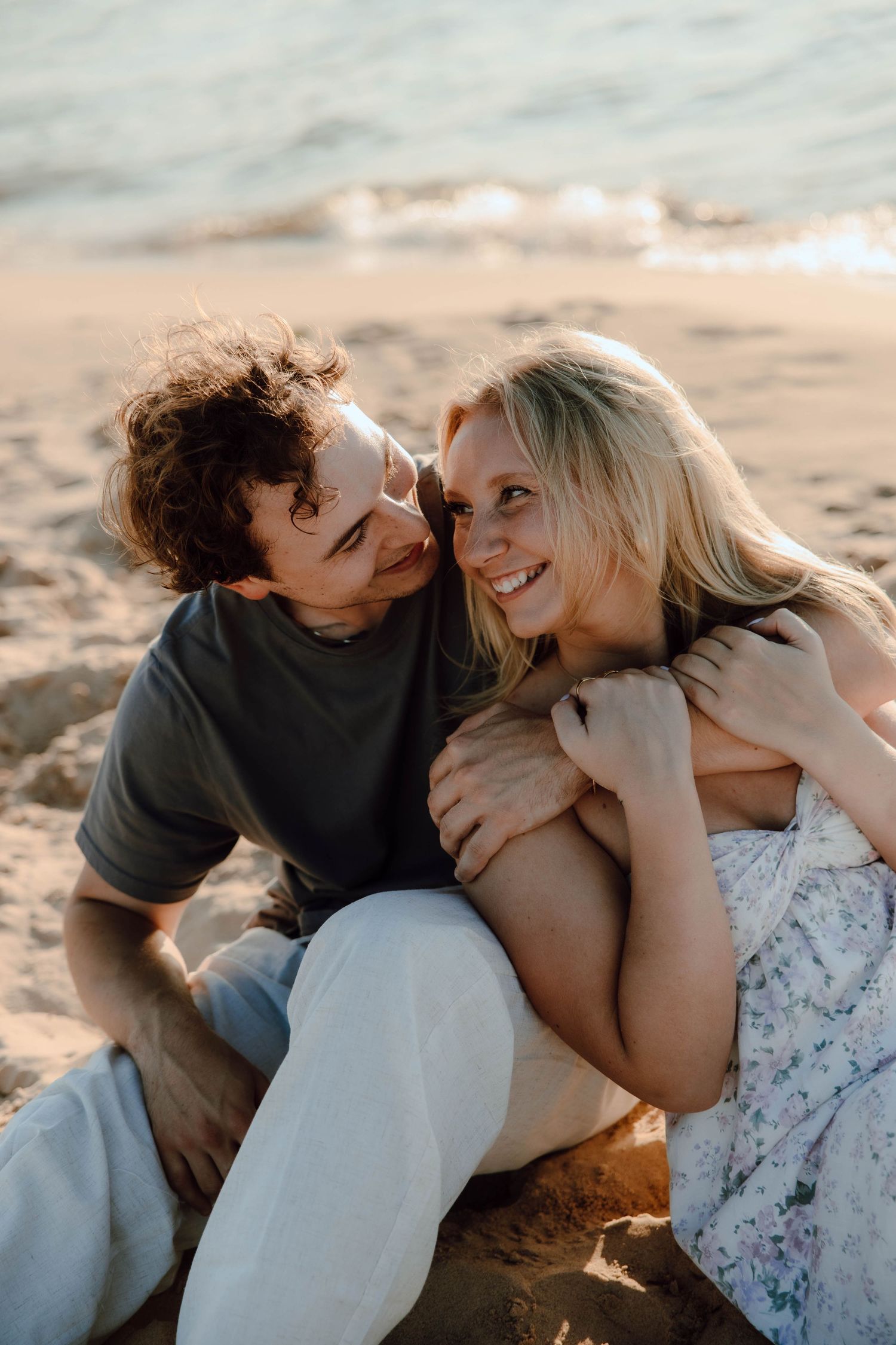 A couple sits on a sandy beach at sunset, smiling and embracing in a romantic and candid moment.