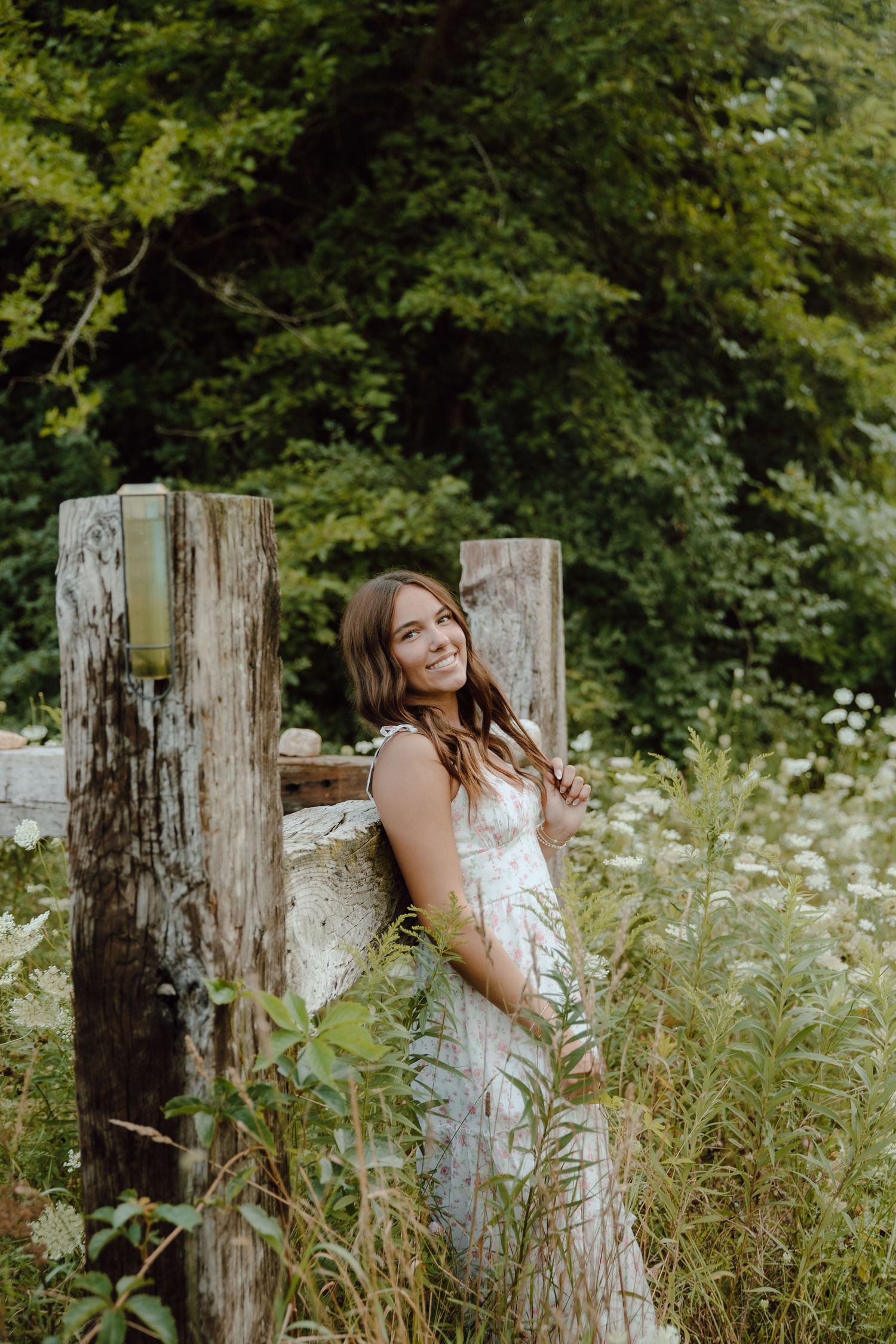 Smiling young woman in white dress leaning against rustic wooden fence posts surrounded by wildflowers and lush greenery.