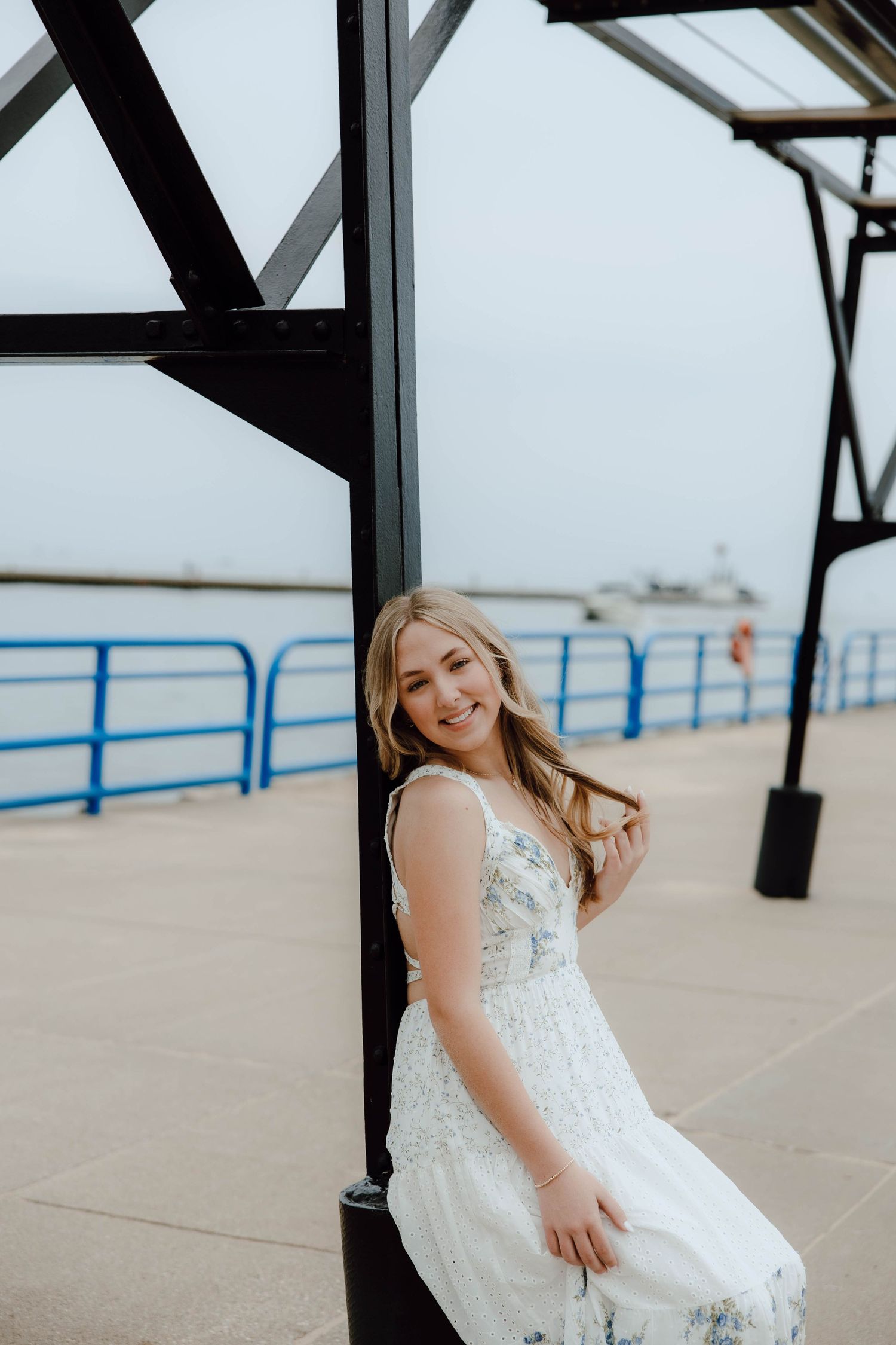 Smiling blonde woman in a floral white dress posing on a waterfront pier with blue railings and overcast skies.