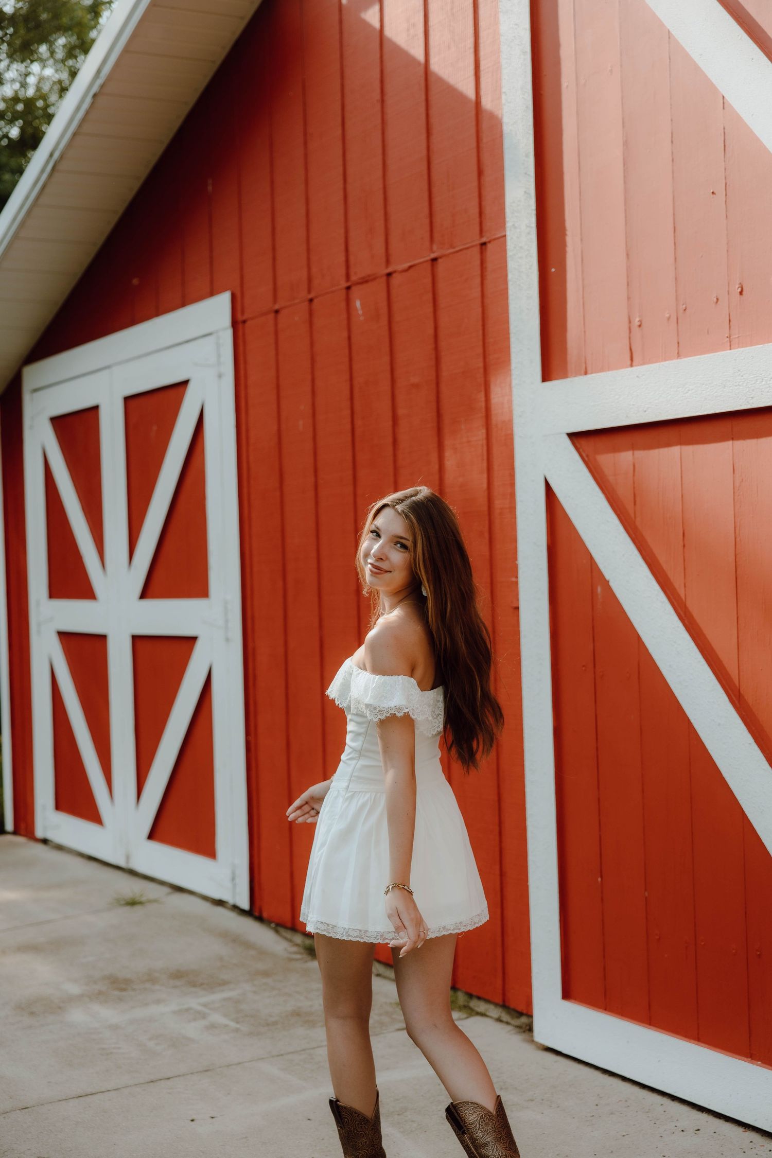 Young woman in white off-shoulder dress and cowboy boots posing in front of a red barn.