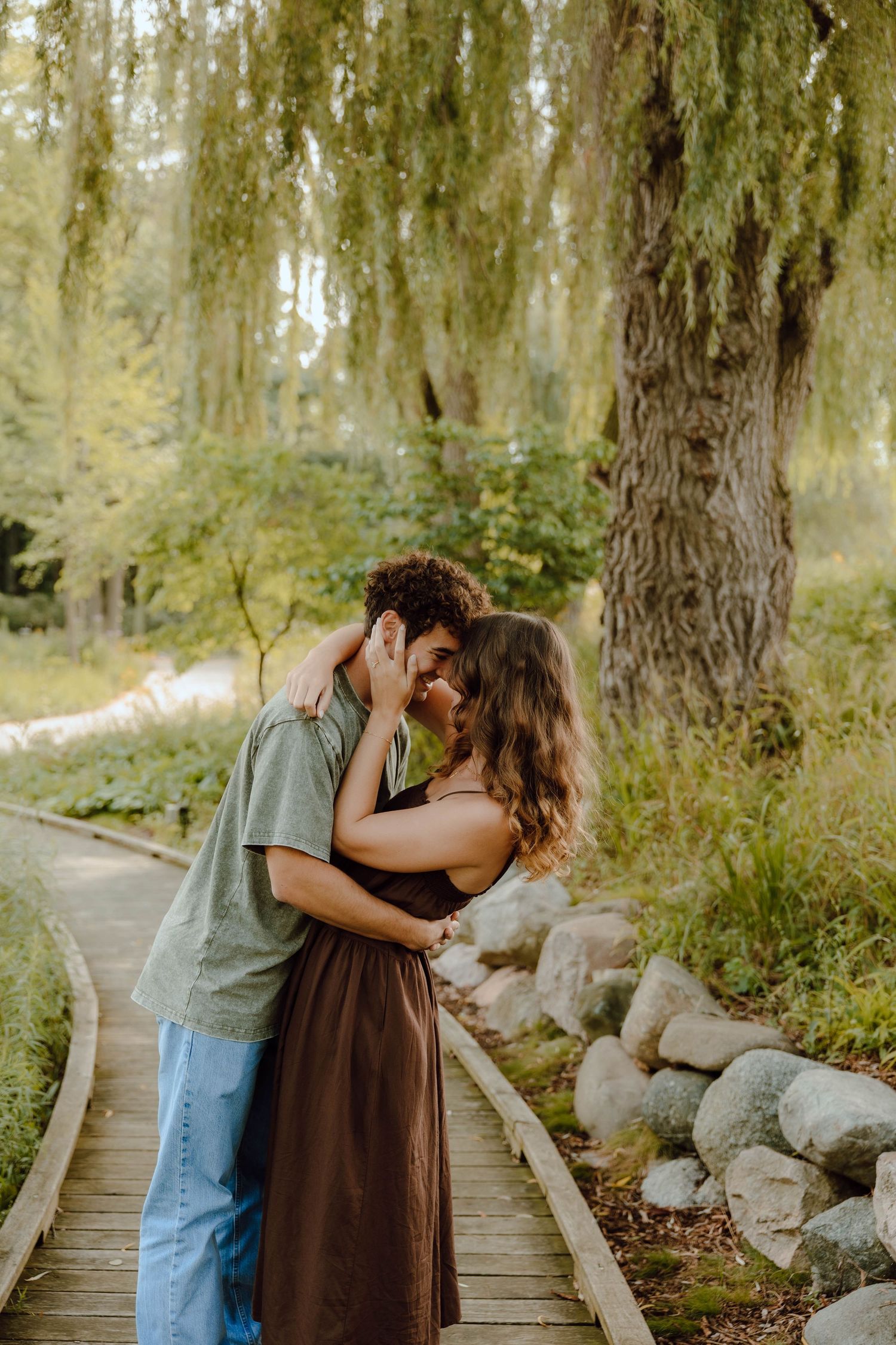 Couple sharing a romantic kiss on a wooden boardwalk path surrounded by lush green willow trees in a park.