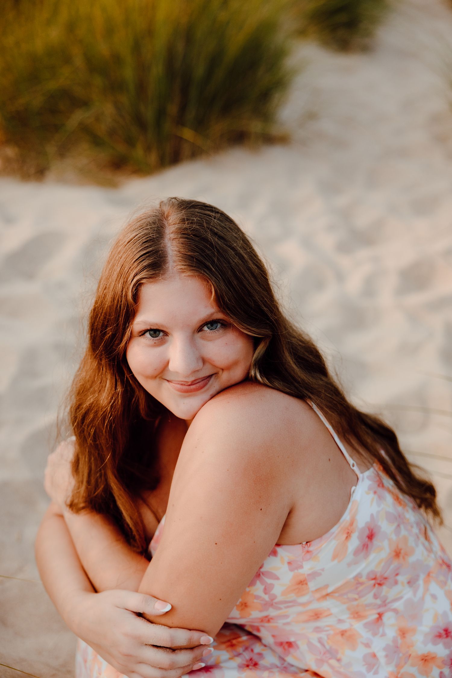 Portrait of a person in a floral dress sitting on sandy beach with a warm smile.