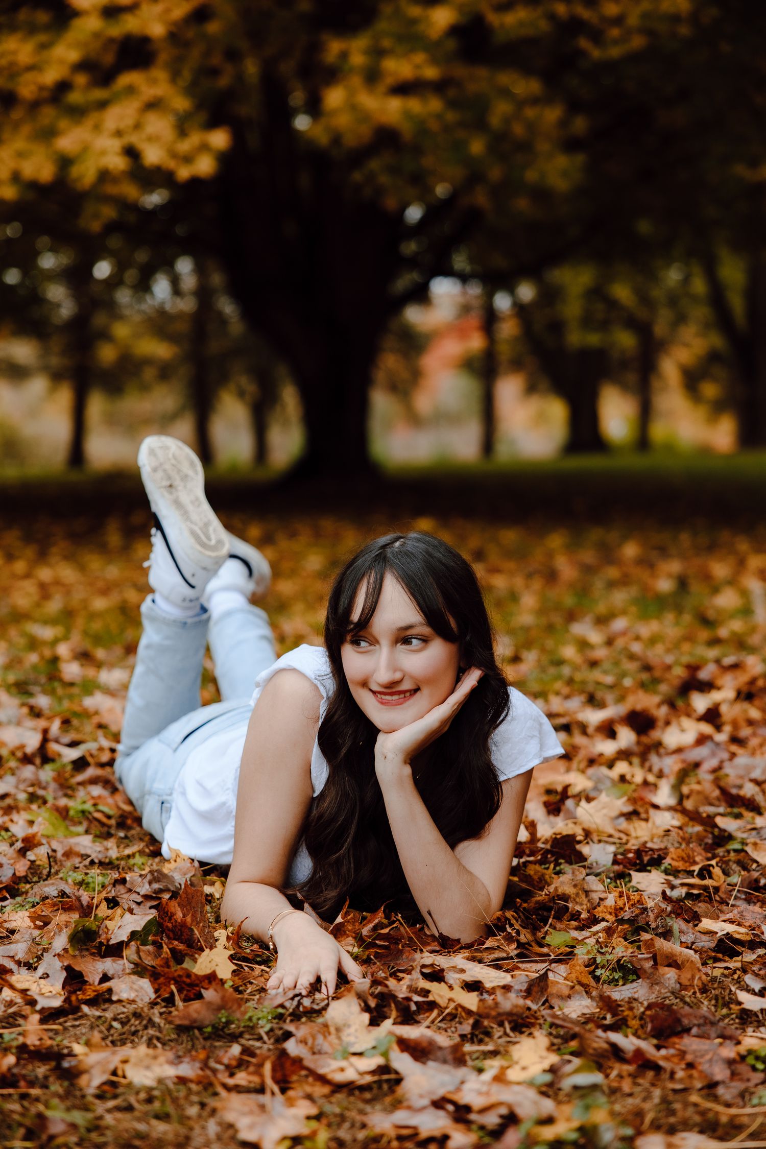 Relaxed pose on autumn leaves wearing white top and light jeans during outdoor photography.