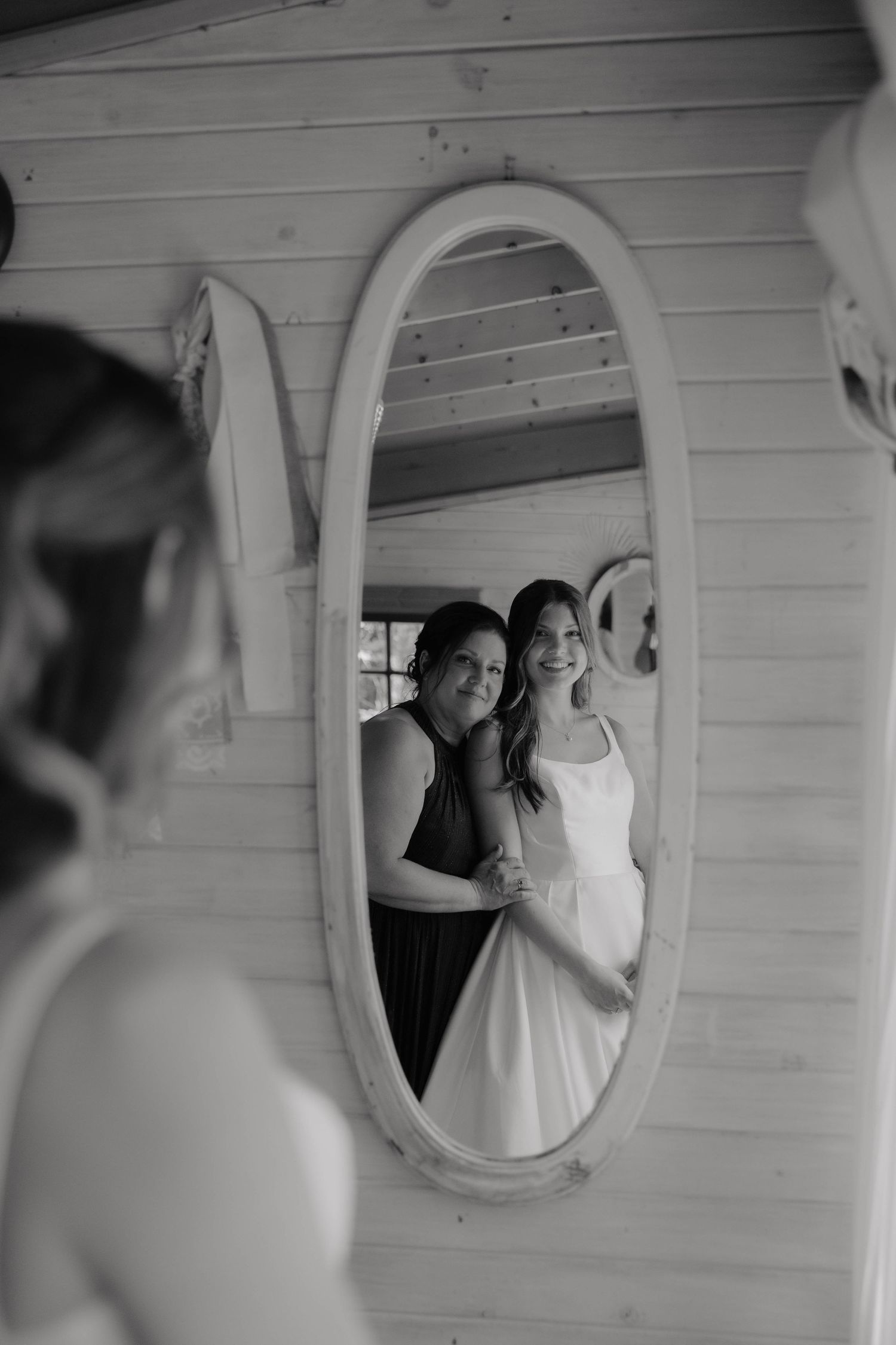 Black and white mirror reflection of bride and bridesmaid smiling together during wedding preparations.