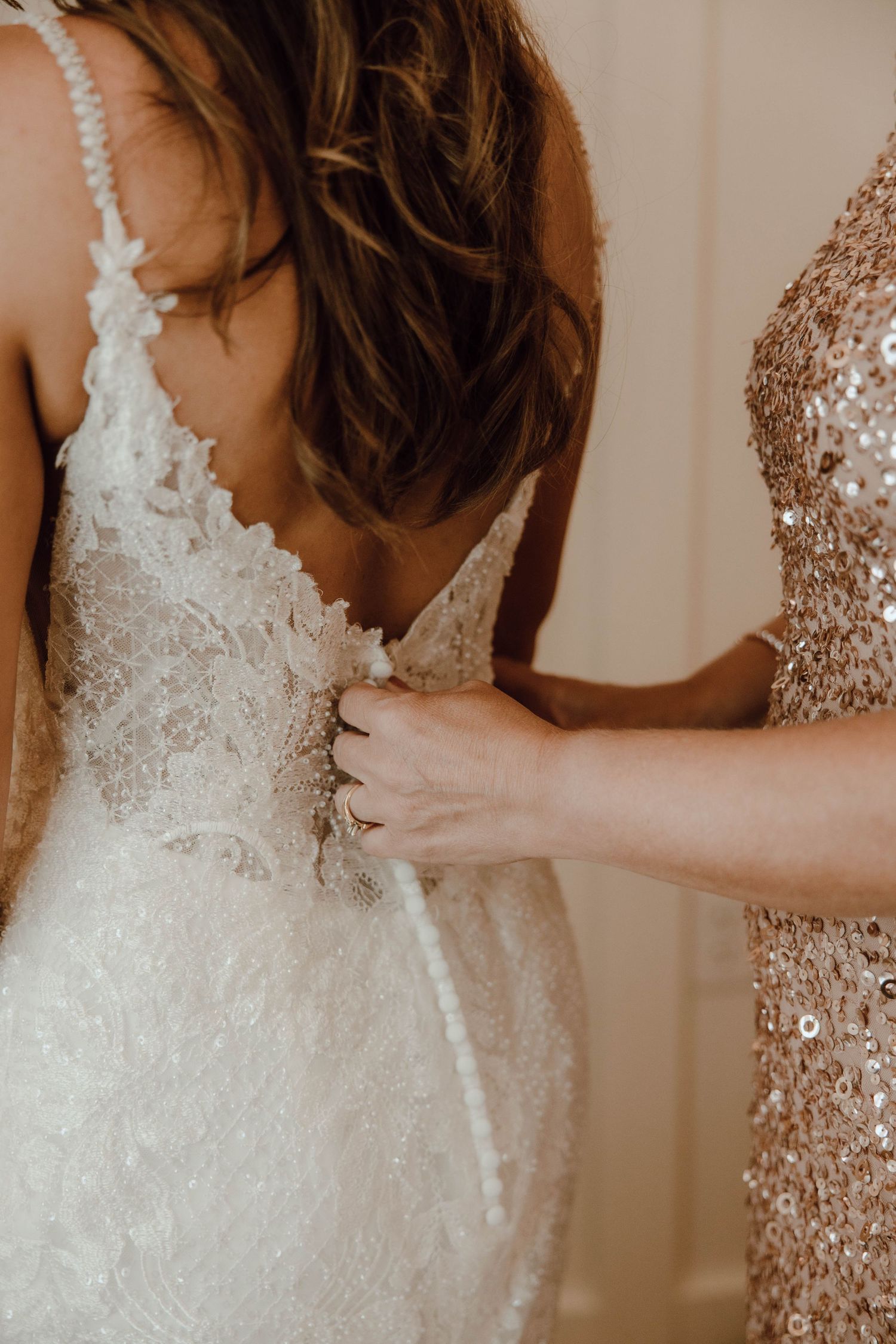 Bridesmaid in sequin dress buttoning the back of a lace wedding gown on the bride.