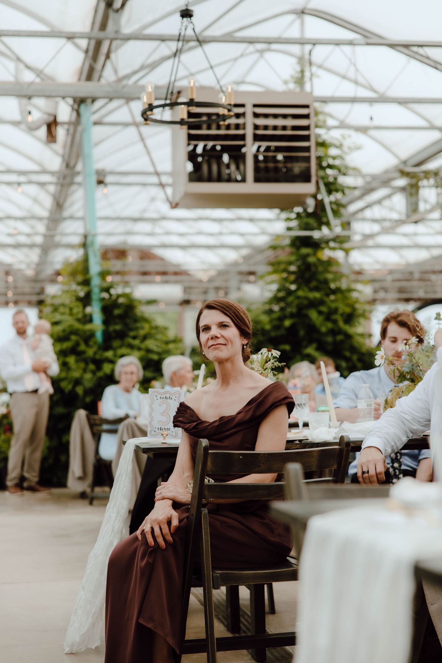 Bridesmaid in burgundy off-shoulder gown seated at table during elegant greenhouse wedding reception.