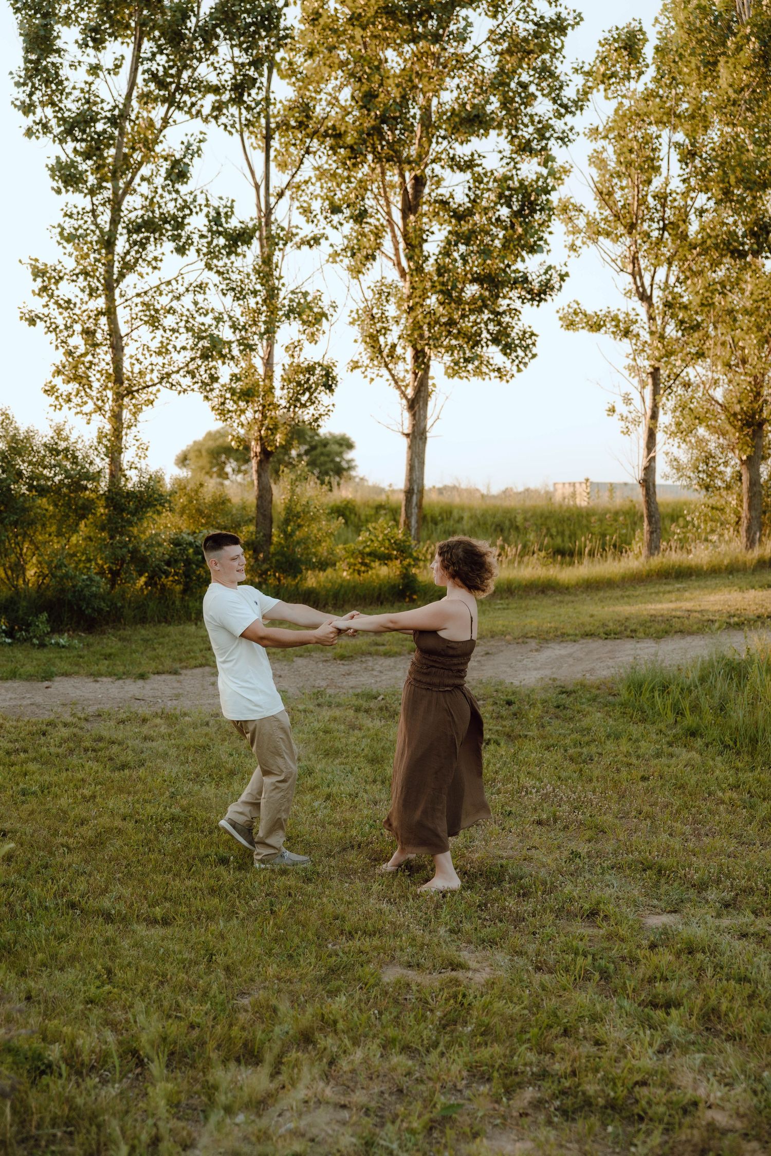 A couple holds hands and spins together in a sunny green field at golden hour, surrounded by tall trees.