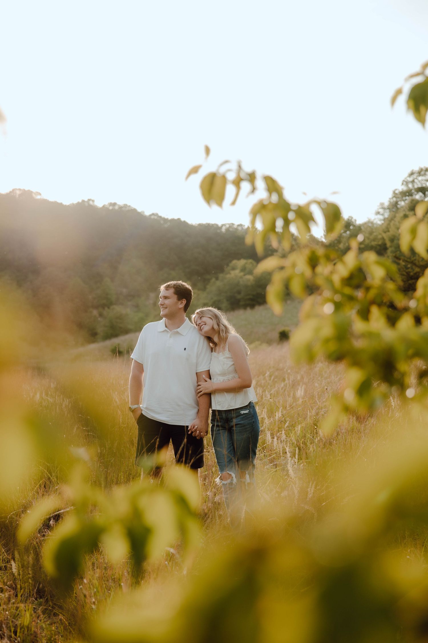 Couple standing in golden sunlit meadow, woman leaning on man in white polo, surrounded by lush green hills.
