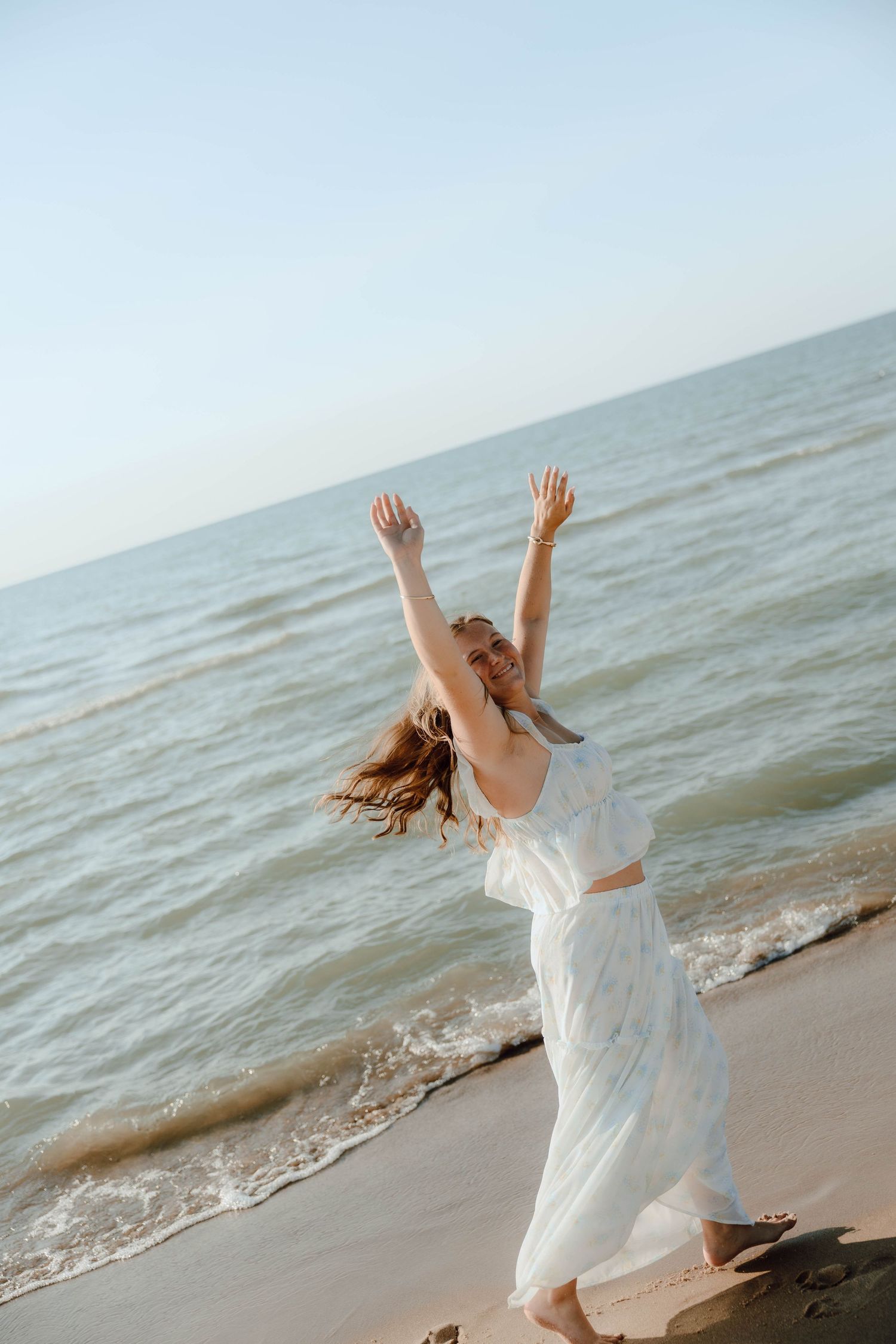Woman in white dress joyfully raises arms on a sunny beach with ocean waves in the background.