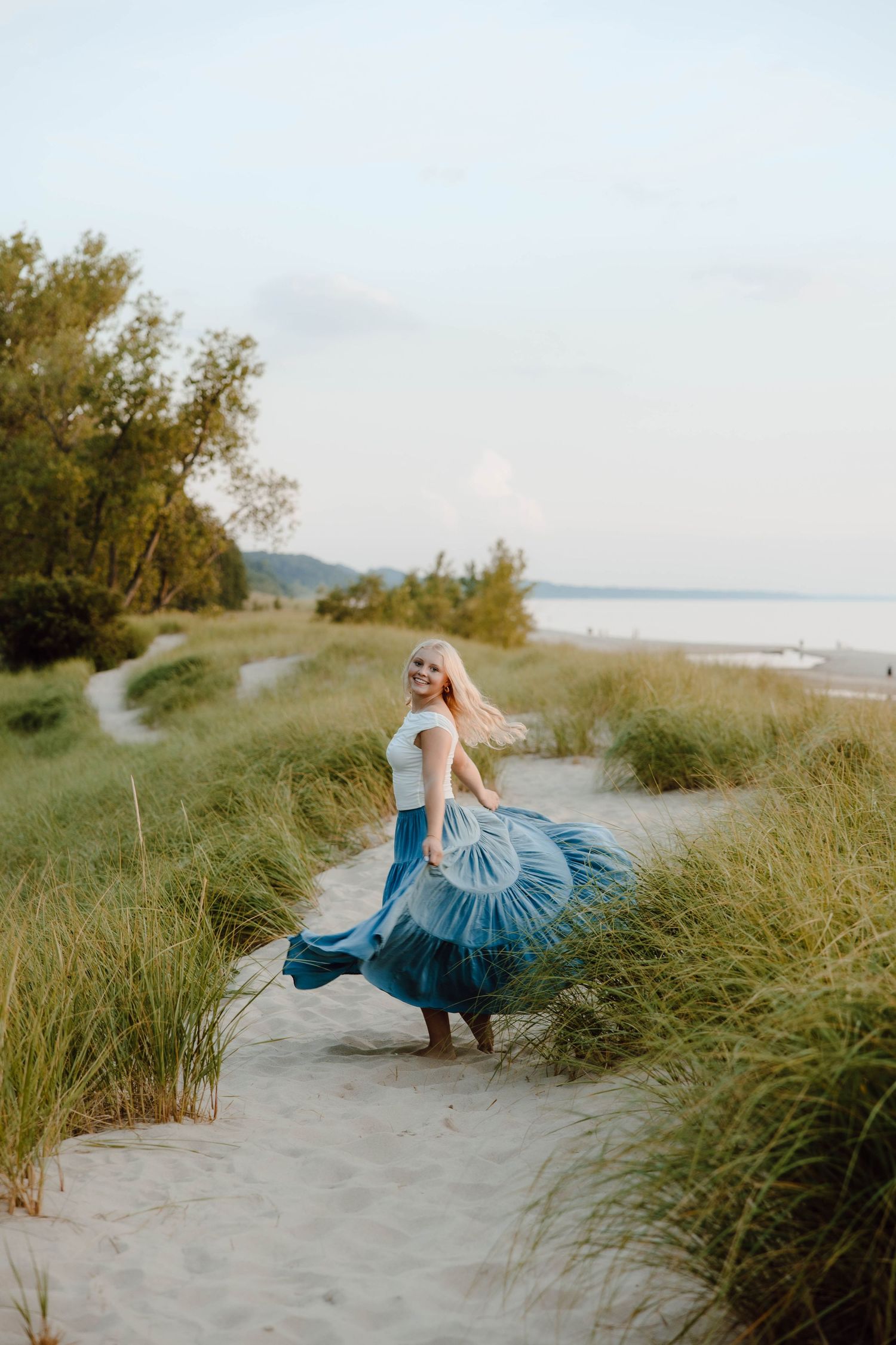 Blonde woman twirling in a blue flowing skirt on a sandy beach path surrounded by green grass at sunset.