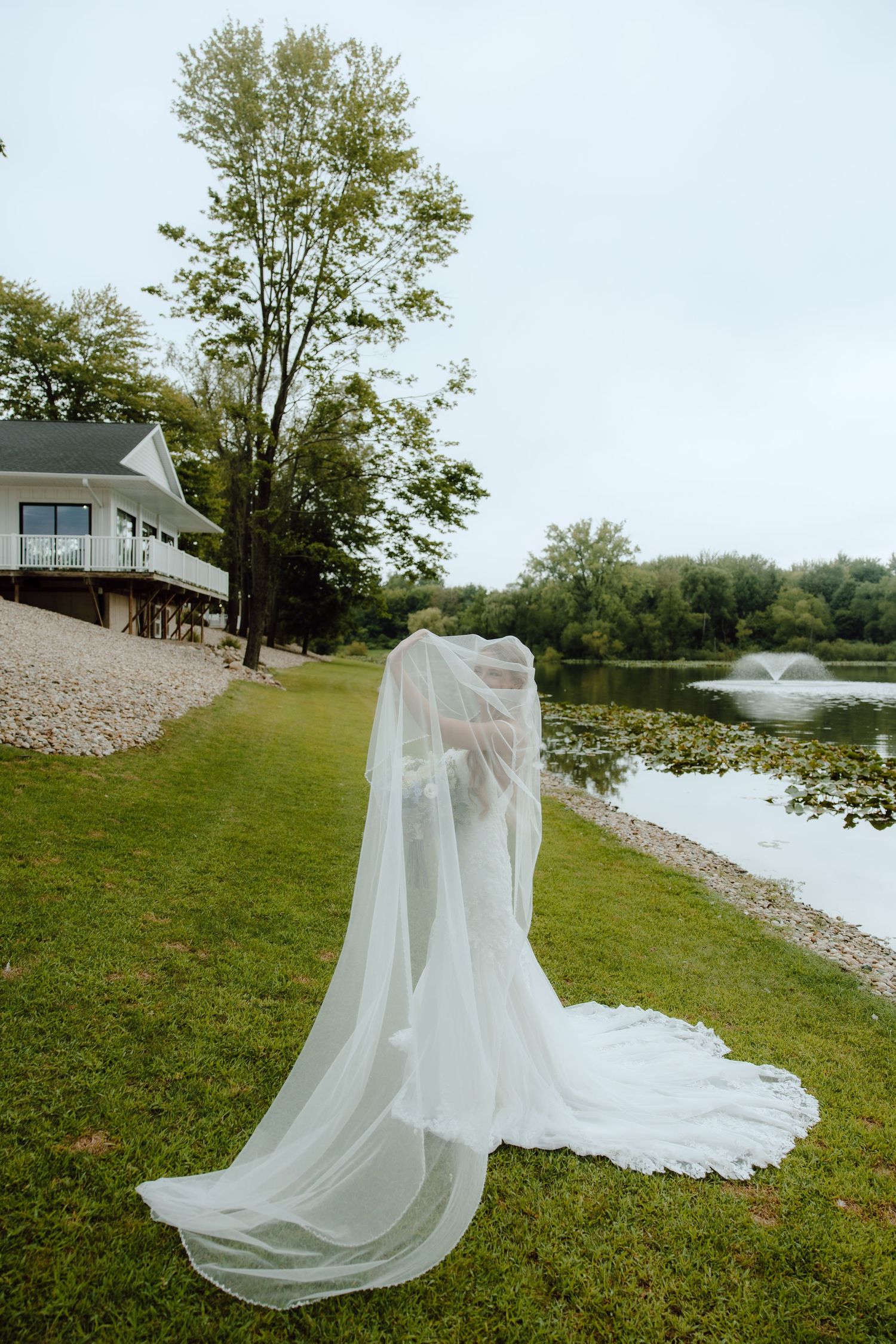 Bride in flowing white wedding gown and veil standing by a serene pond with lush greenery and a white house backdrop.