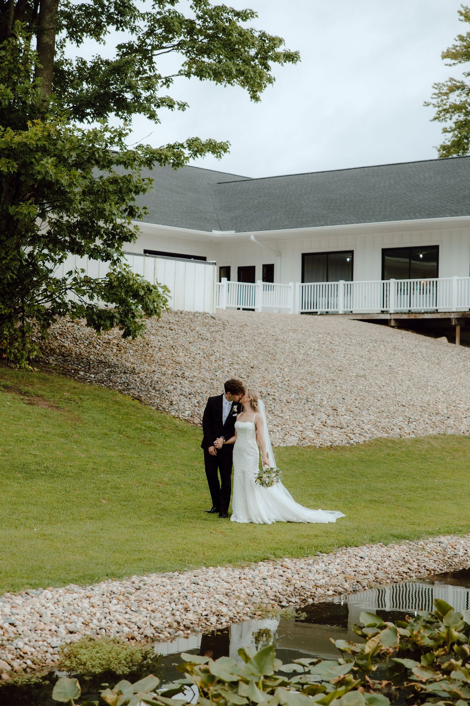 Bride and groom sharing kiss near pond outside white wedding venue surrounded by greenery.