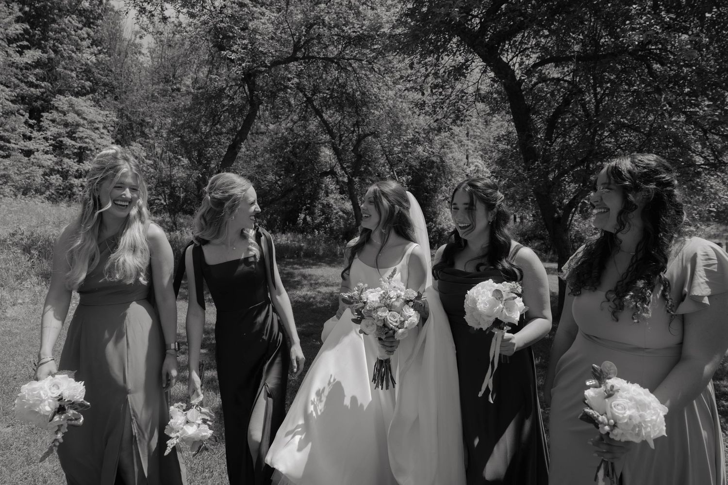 Bride in white gown with five bridesmaids in dark dresses holding bouquets outdoors in black and white photo.