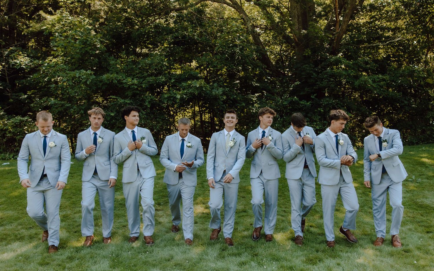 Nine groomsmen in matching light blue suits pose playfully on green lawn with trees behind.
