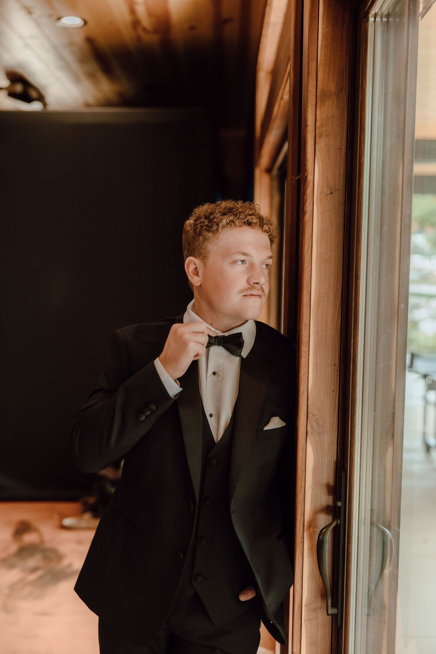 A groom in a black tuxedo with bow tie adjusts his collar while standing by a wooden door frame.