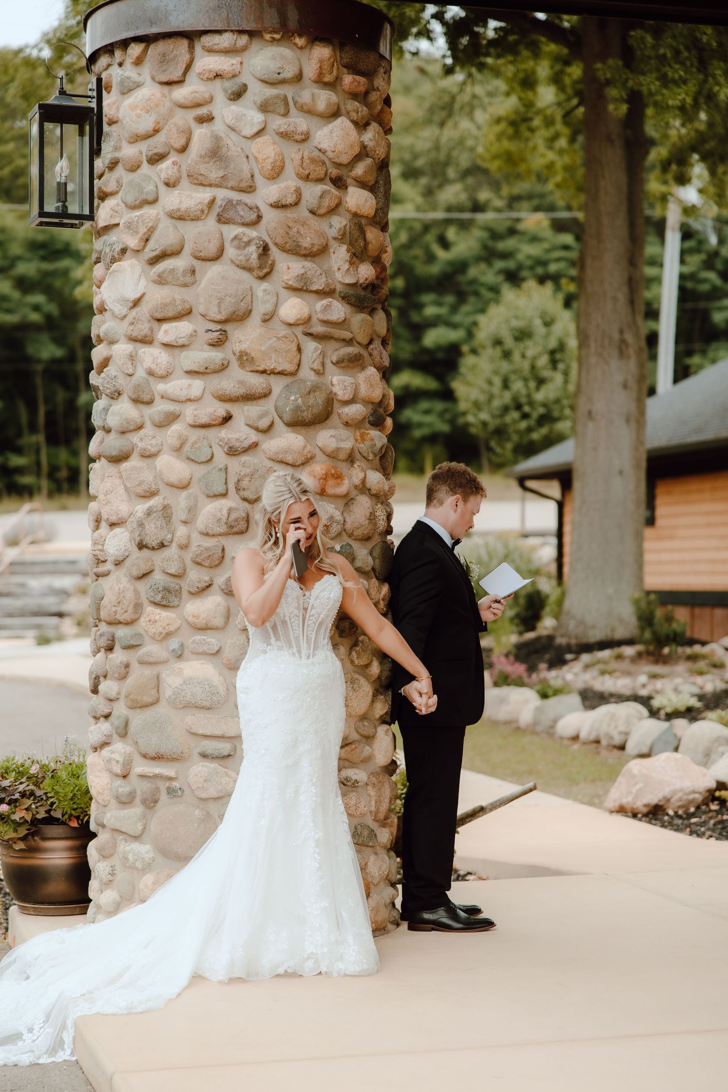 Bride and groom share private vows beside a stone pillar at an outdoor wedding venue surrounded by lush greenery.