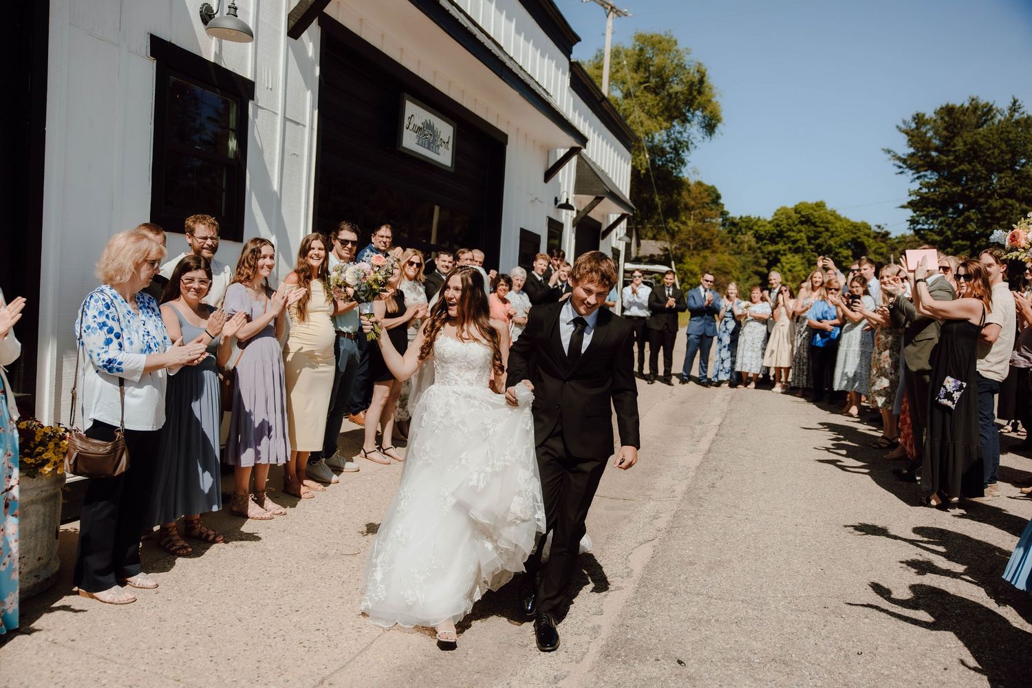 Bride and groom walk joyfully through cheering wedding guests outside a rustic venue on a sunny day.
