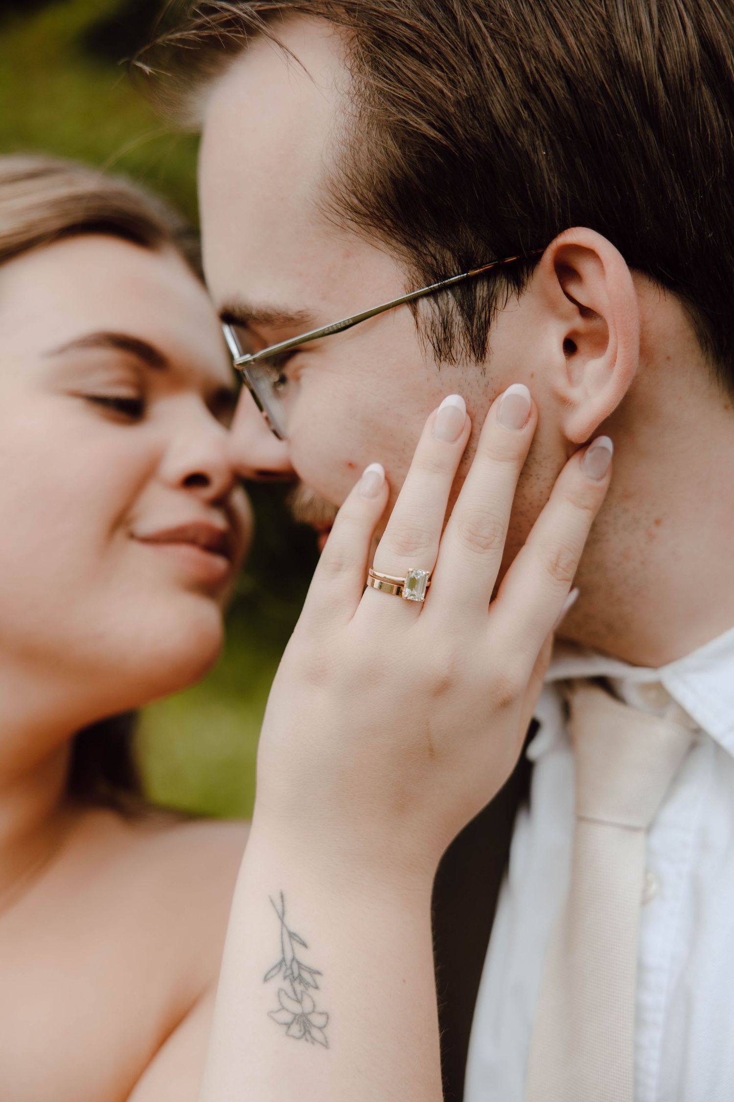 Close-up of couple almost kissing, woman showing engagement ring with floral wrist tattoo, man wearing glasses.