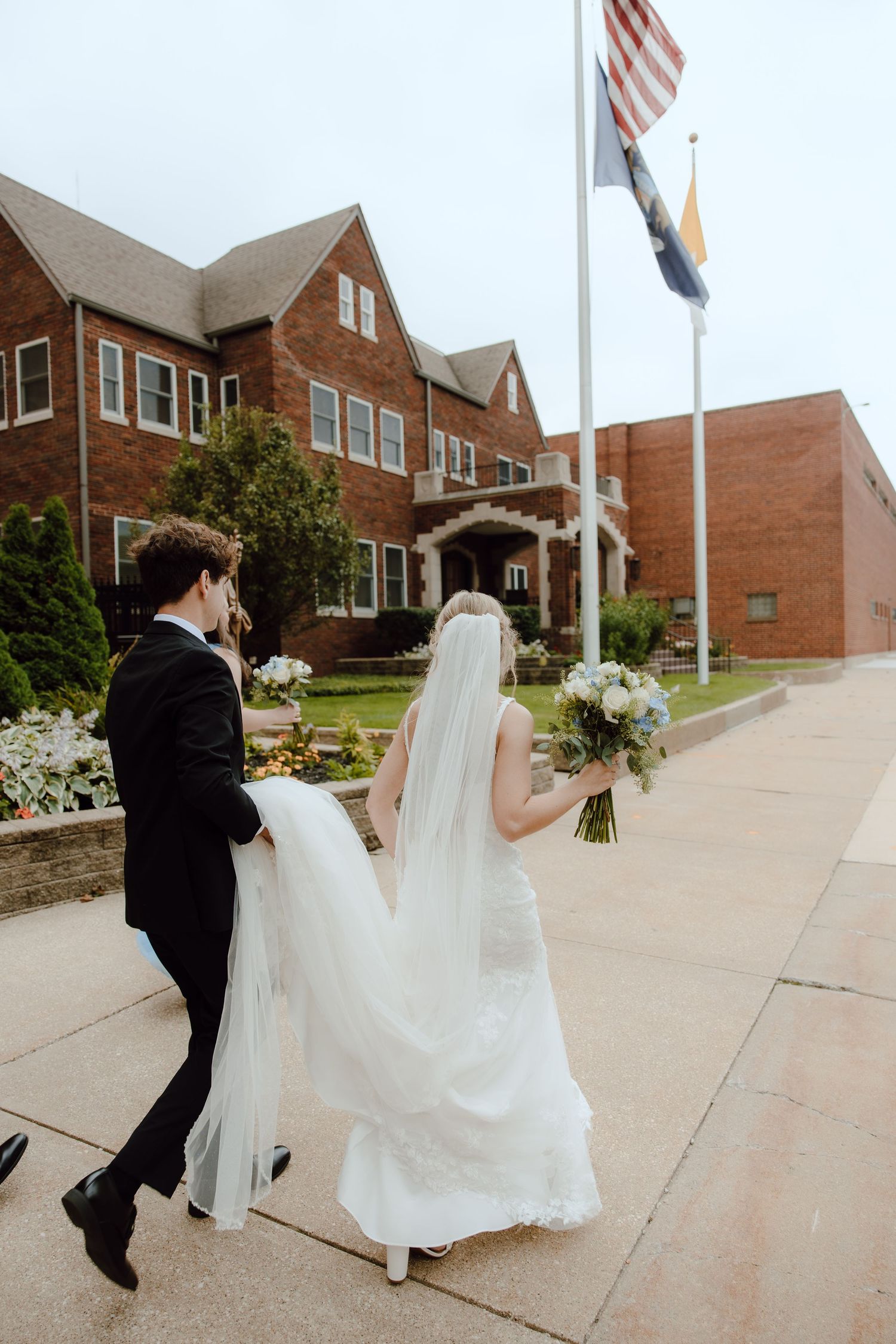 Bride and groom walk outside brick church building, groom carrying wedding dress train.