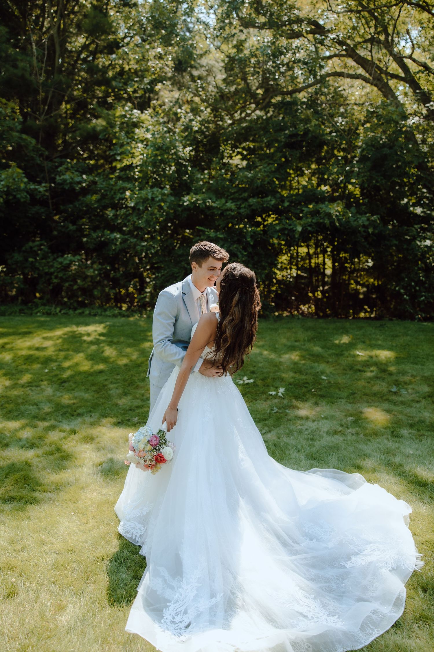 A bride in a flowing white gown holds a bouquet while sharing a romantic moment with her groom outdoors.