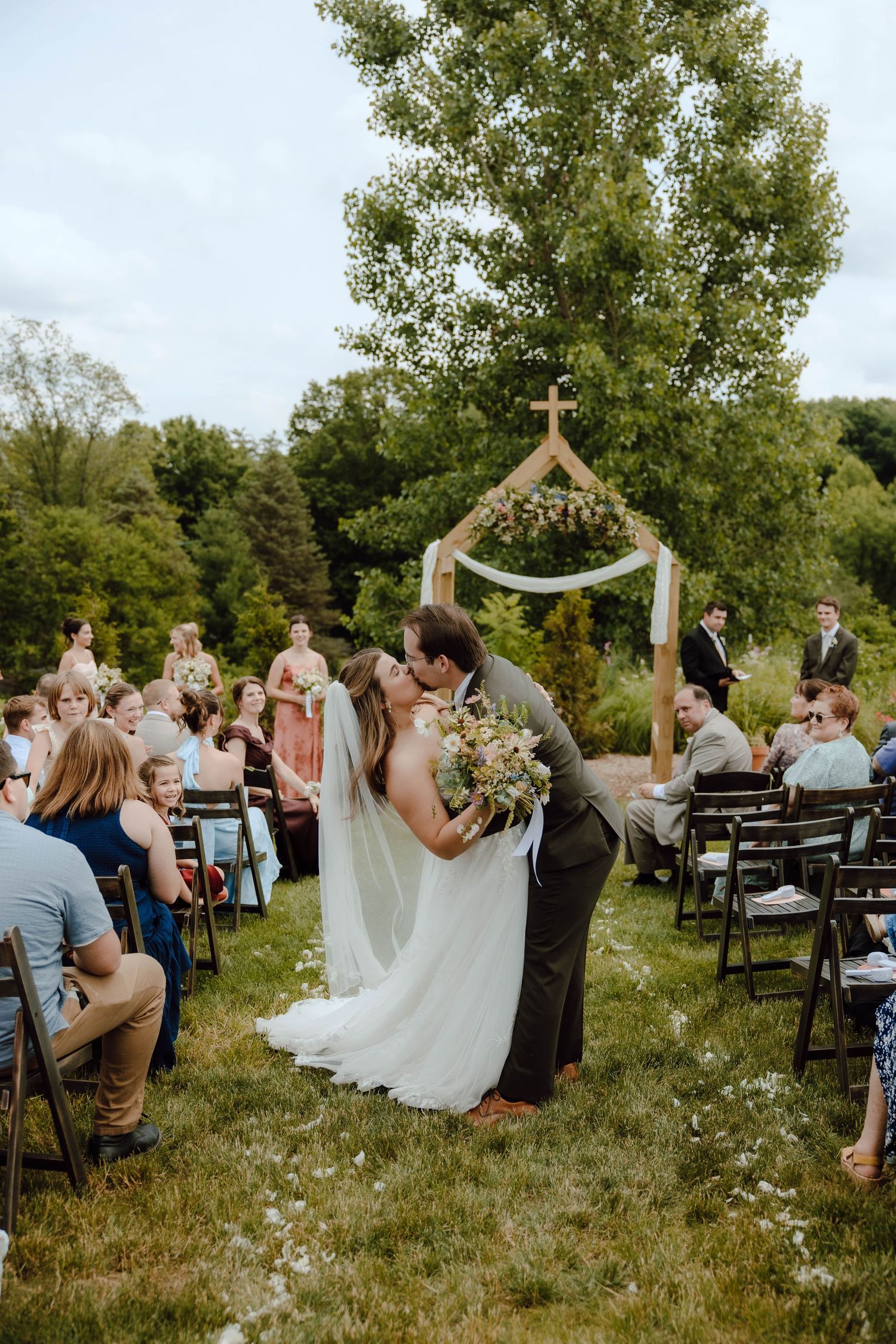 Bride and groom sharing first kiss at outdoor wedding ceremony with wooden cross arch.