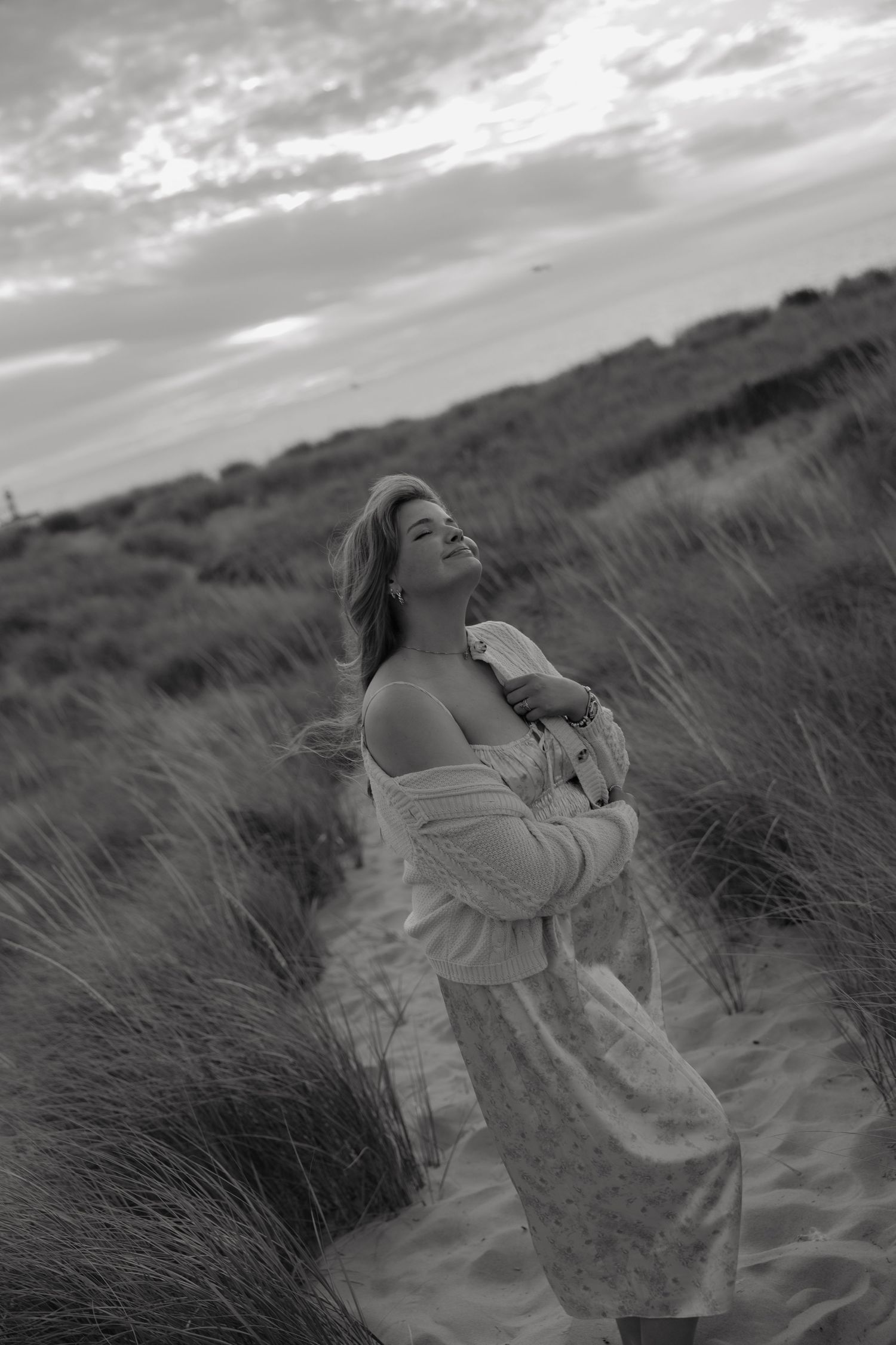 Black and white photo of a woman in a flowy dress standing among beach dunes, head tilted back with eyes closed.