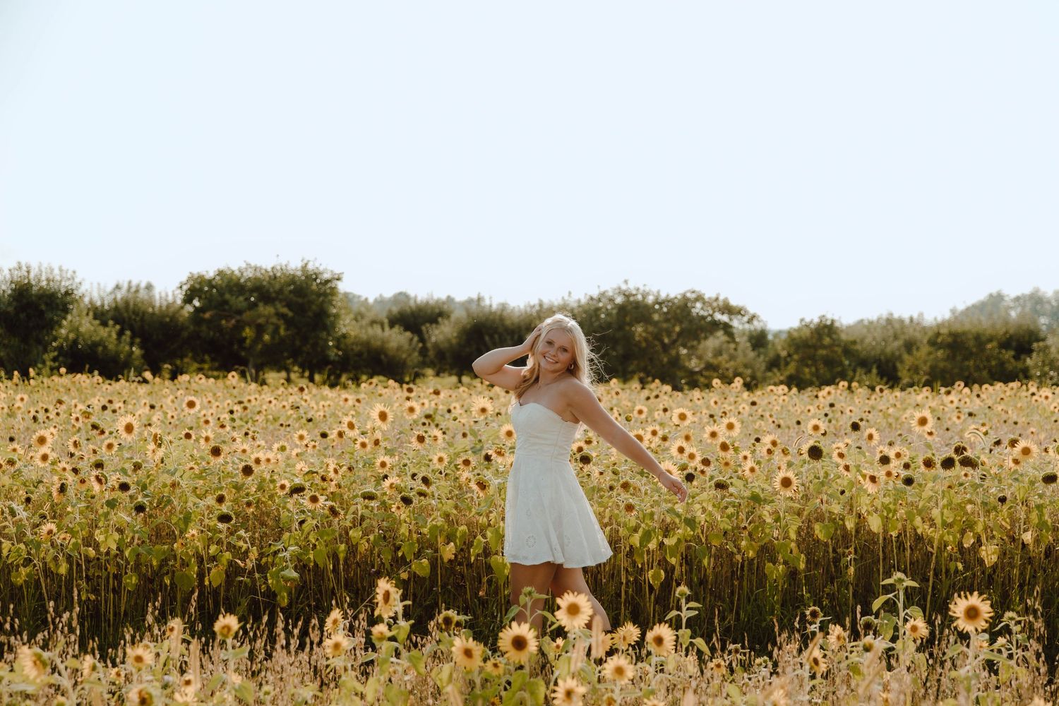 A young blonde woman in a white dress dancing joyfully in a vibrant sunflower field on a sunny day.