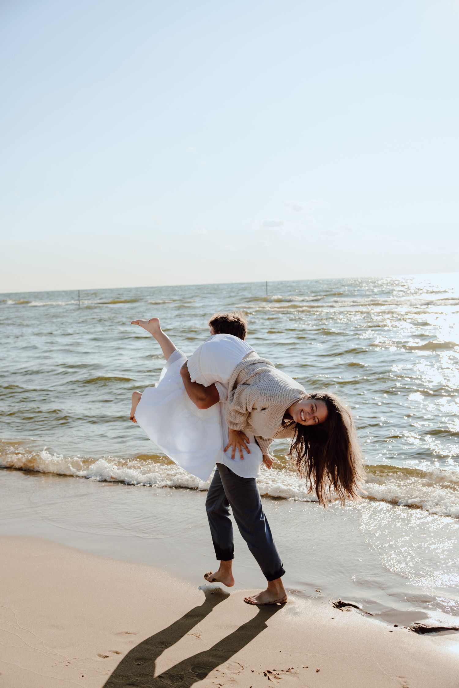 A person dips their partner back playfully on a beach with ocean waves behind them.