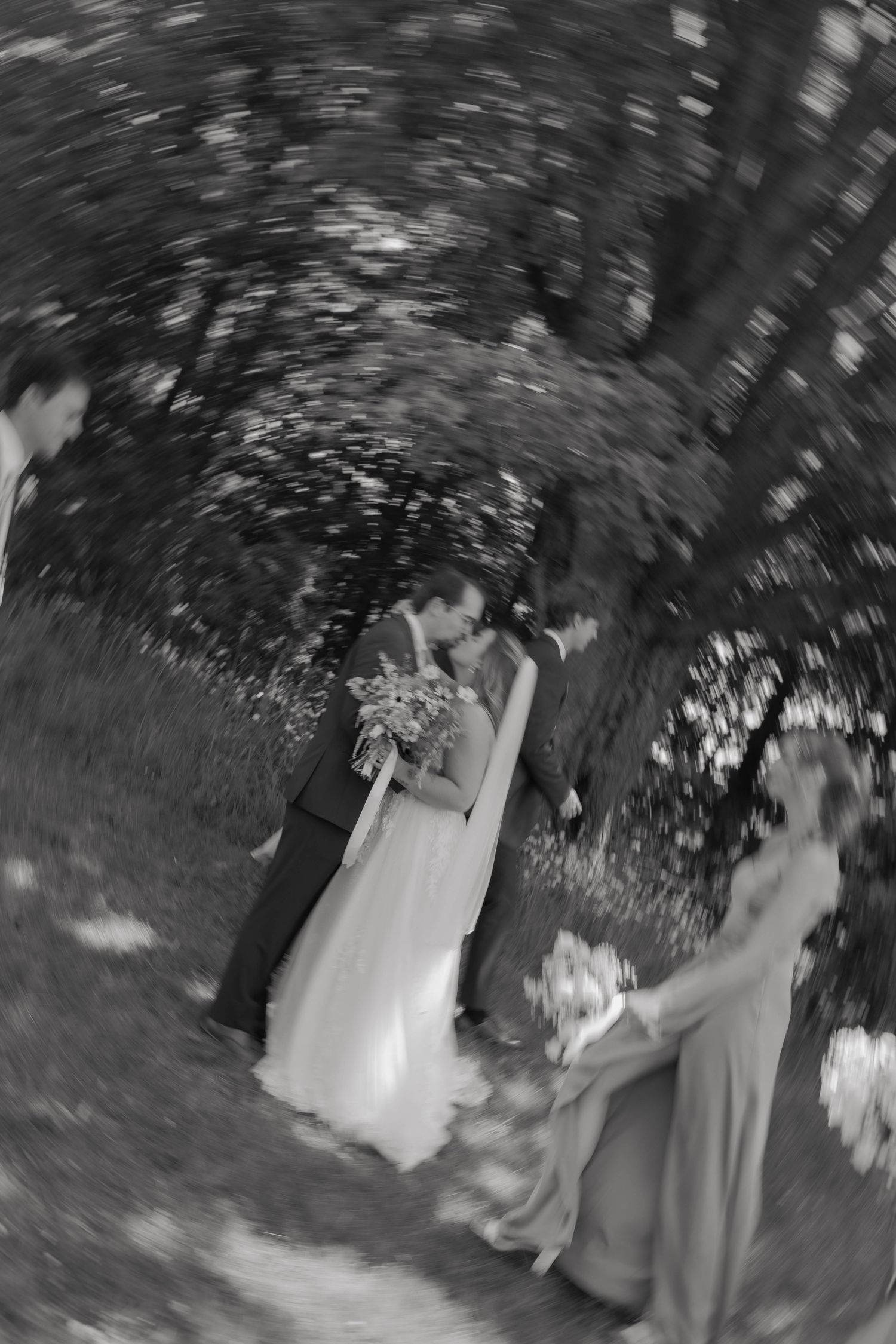 Black and white fisheye lens photo of a bride and groom kissing under trees.
