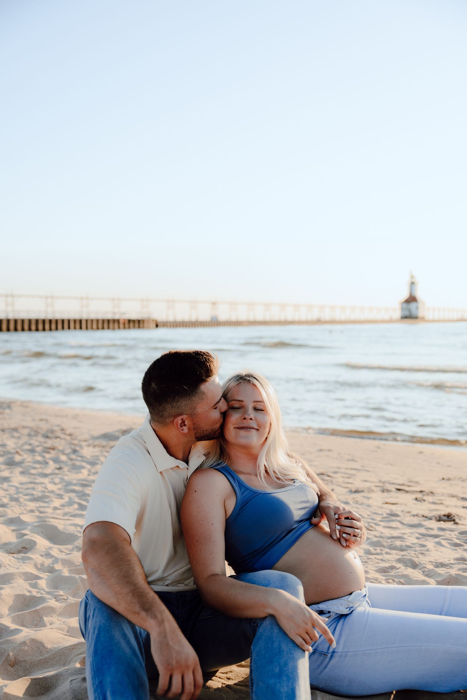 Romantic beach maternity photos with lighthouse pier in background at sunset.