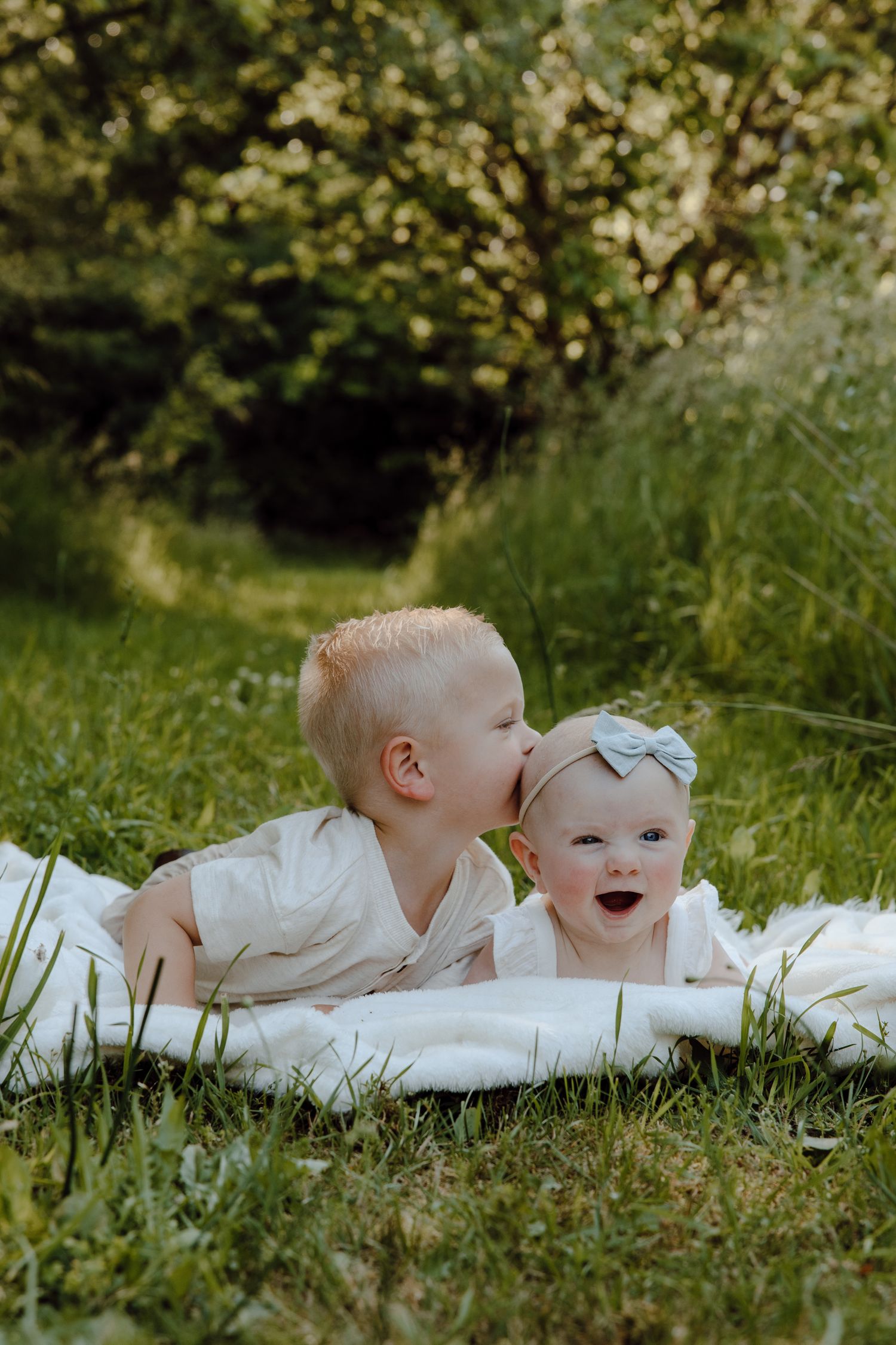 Two young children in white outfits lying on a blanket in green grass.