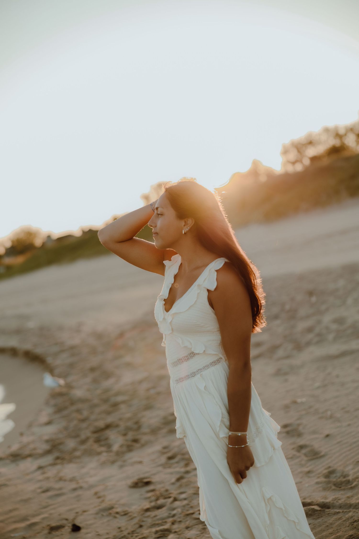Woman in white dress on sandy beach at golden sunset, hand raised to head, backlit by warm sunlight.