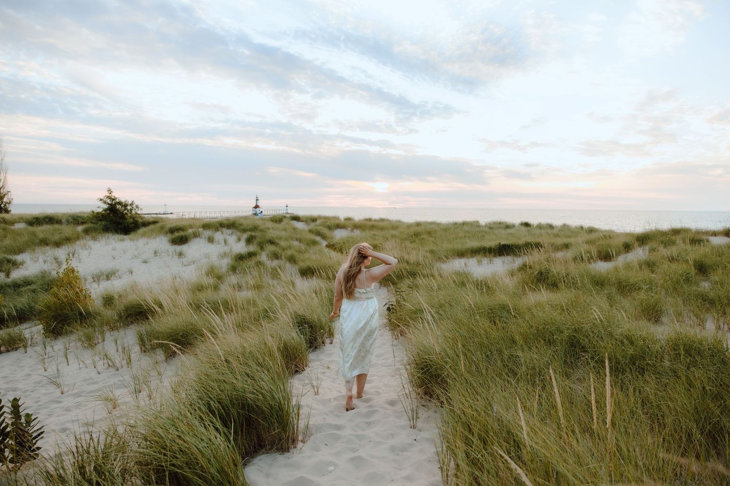 A woman in a light dress walks barefoot through sandy beach dunes with tall grass under a pastel evening sky.